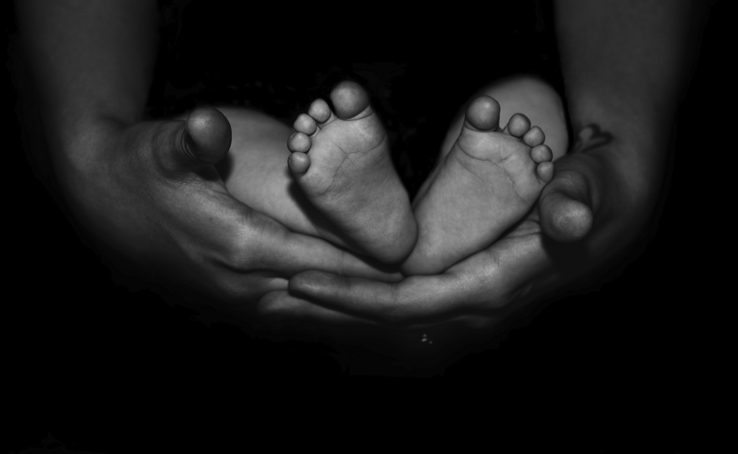 Close-up of a woman’s hands holding an infant’s feet. The soles of the infant’s feet are facing the camera and they are cradled in the woman’s palms.