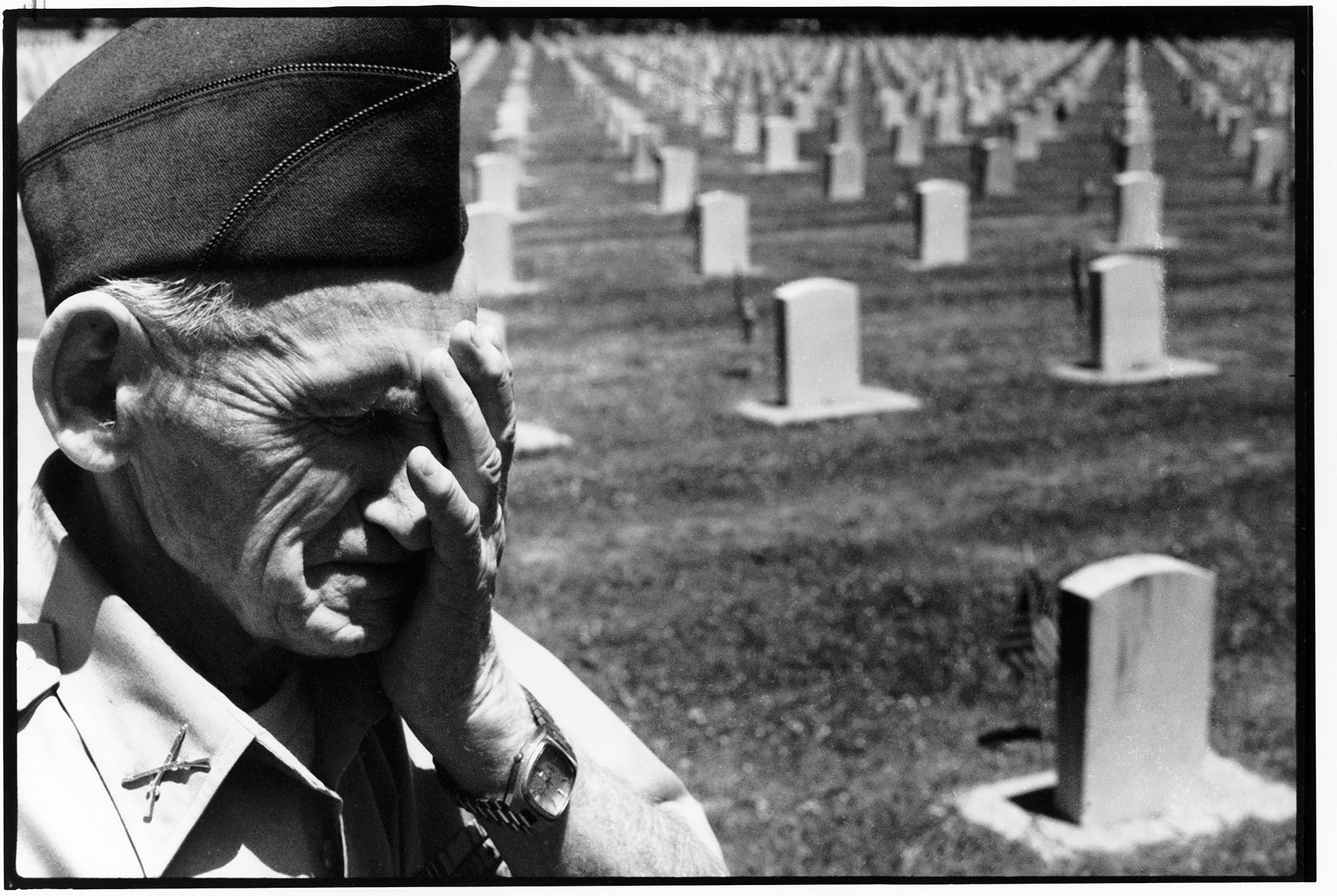 An old soldier in uniform with a crossed-rifles pin on his right lapel stands in a cemetery with his left hand over his face. He looks sorrowful as he stands among the rows of upright headstones, each with a small U.S. flag in front of it.