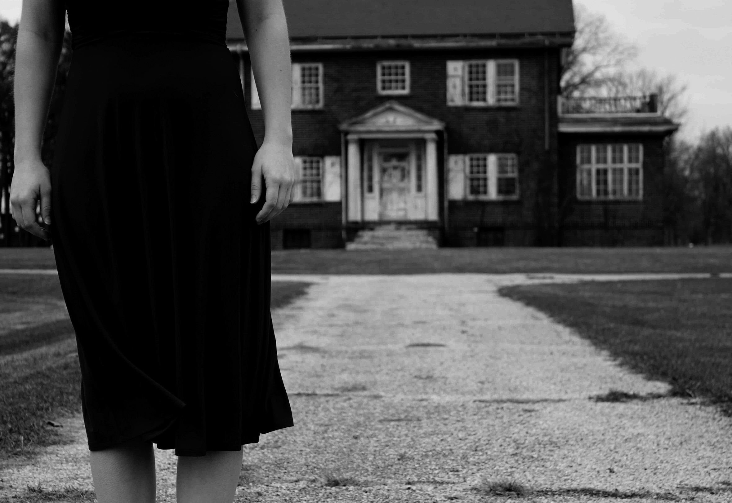 A woman in a dress and facing the camera is seen from the midriff to below the knee with her arms straight down. She is standing on a sidewalk at a distance from the front of an old two-story house.