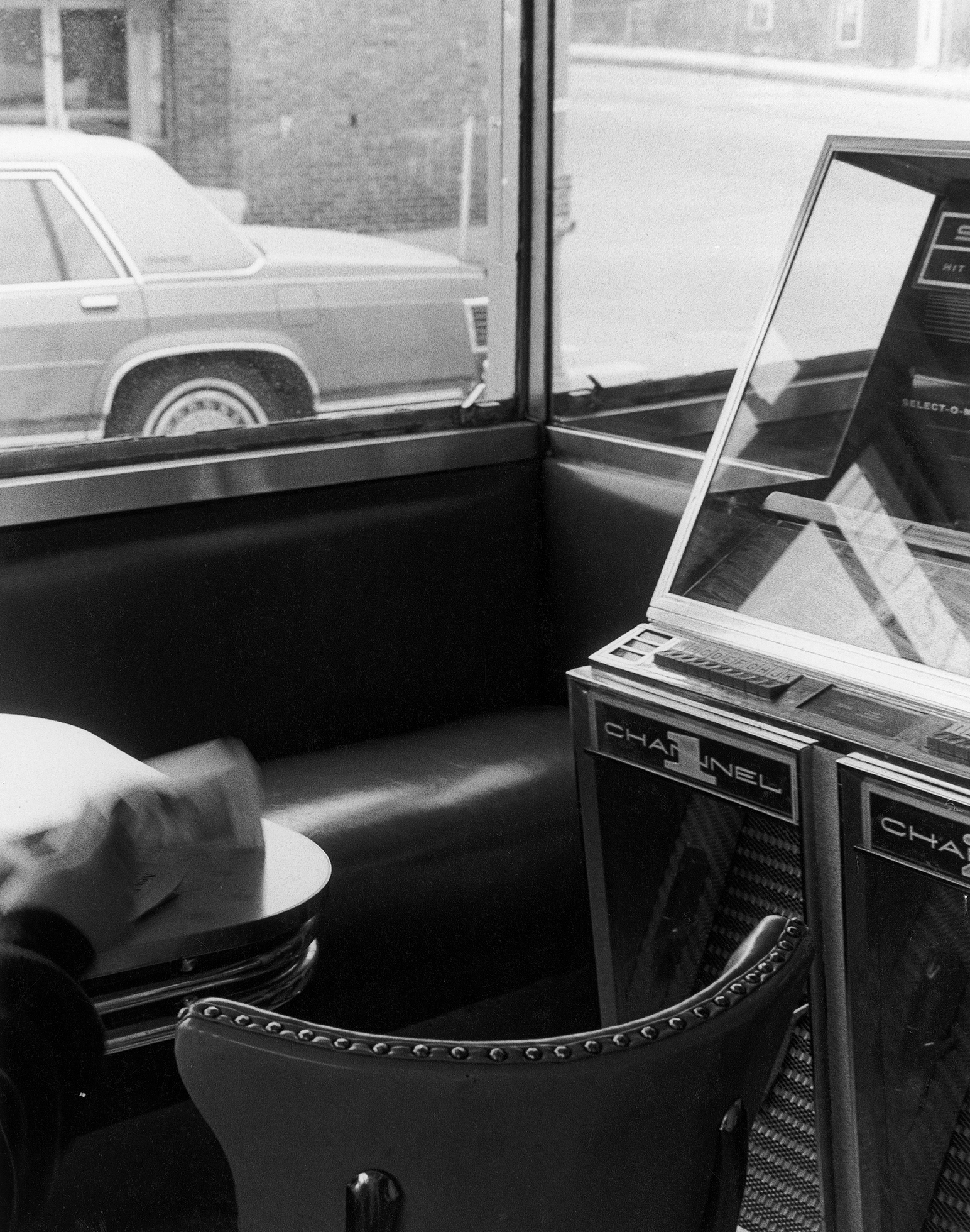 The arm of someone reading the newspaper at a table at a business with bench seating beneath the window and a jukebox. Through the window is a car and brick buildings.
