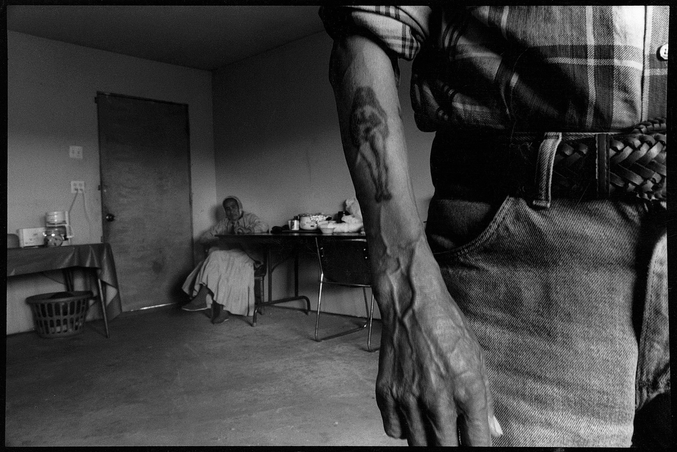 An older woman in a long dress sits in the back corner of a sparsely furnished room while a man stands in the foreground wearing jeans and a plaid shirt with the sleeves folded up. He has a tattoo of a naked woman on his forearm.