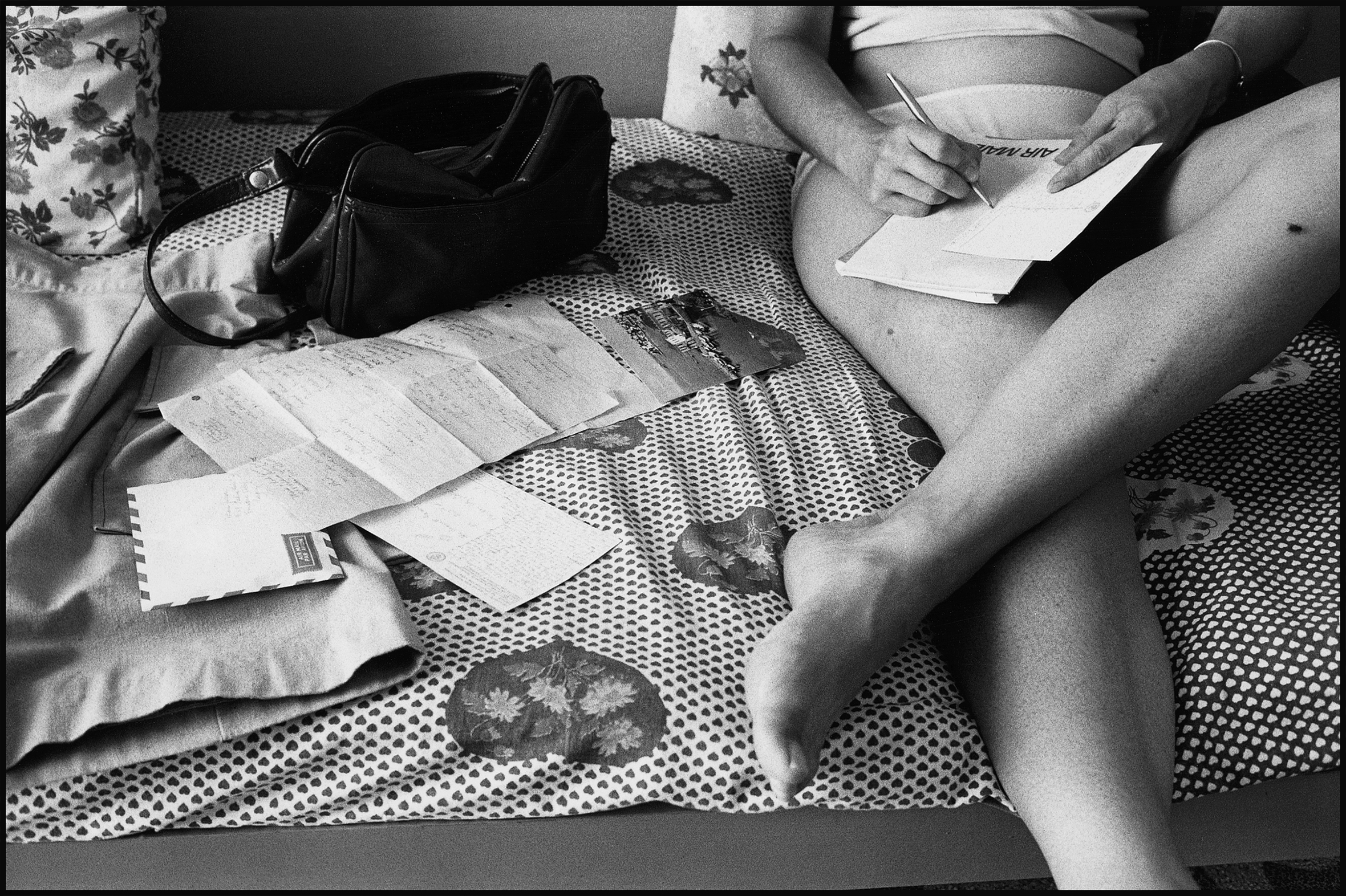 A woman is sitting on a bed with a pillow behind her against the wall writing a postcard on her lap; only the bottom half of her is visible. More postcards and letters are strewn over the bed along with a purse.