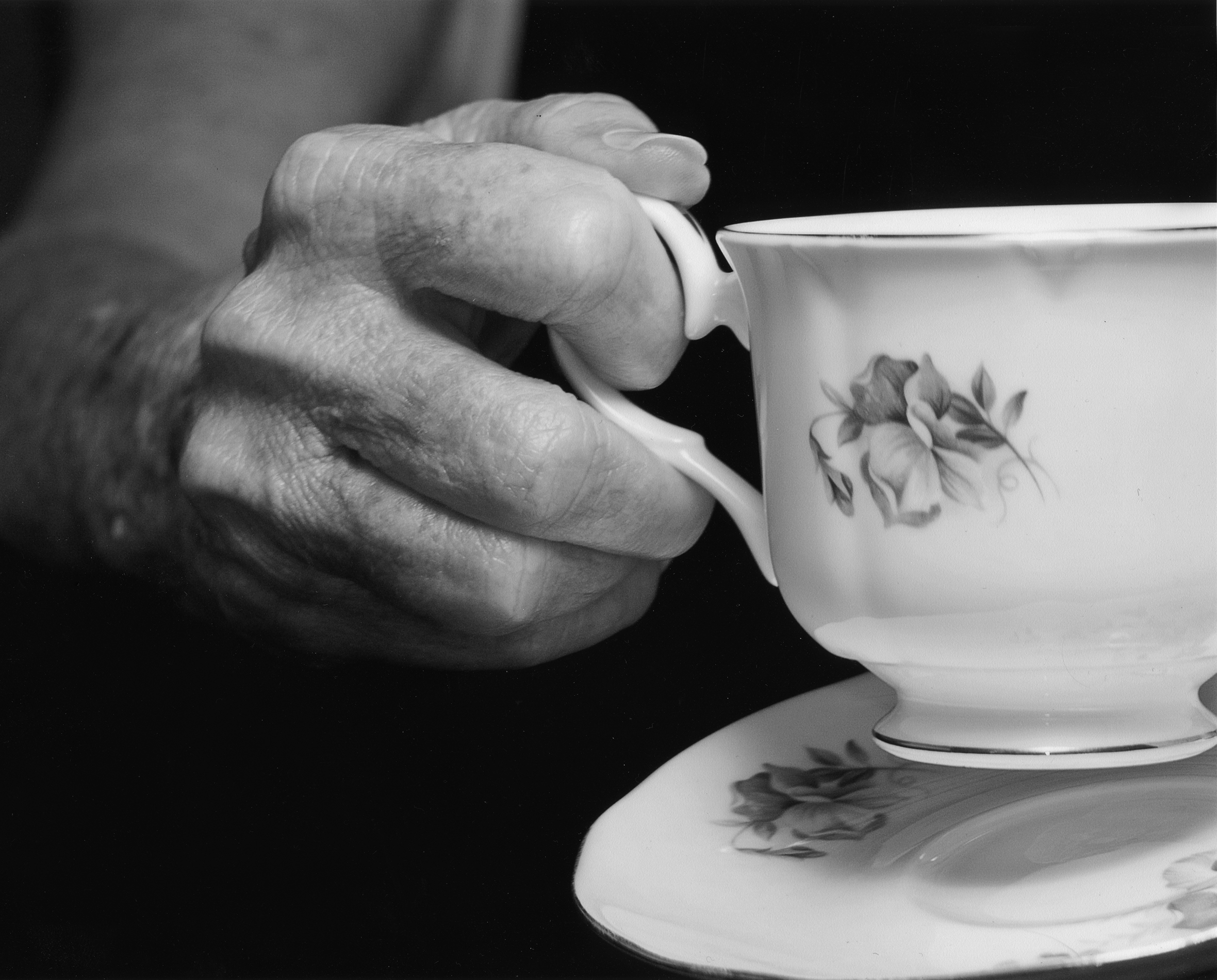 Close-up of the hand of an older woman lifting a flowered teacup by the handle off of its matching saucer.