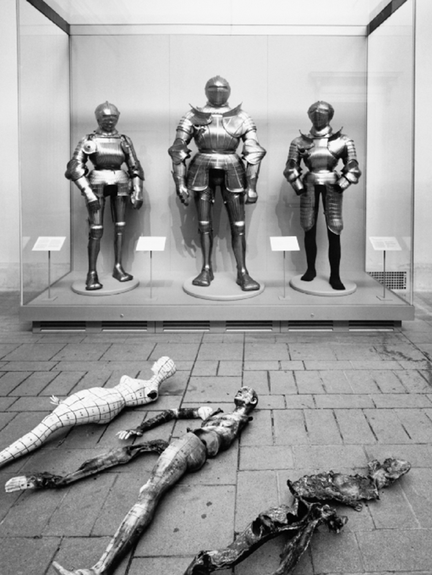 A large display case with three suits of armor standing next to each other. In front of the case on the tile floor are broken mannequins with missing arms and legs.