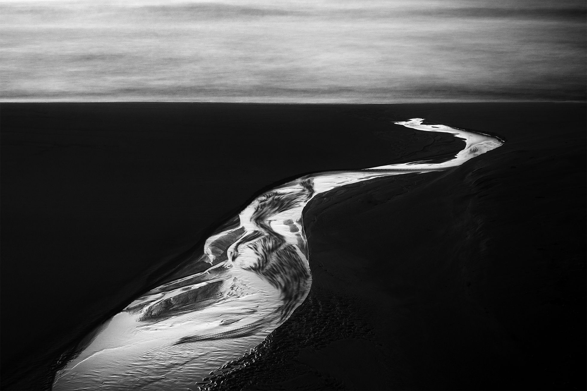 A curving river flowing into the horizon. The water shines and shows movement within the dark landscape on both sides.