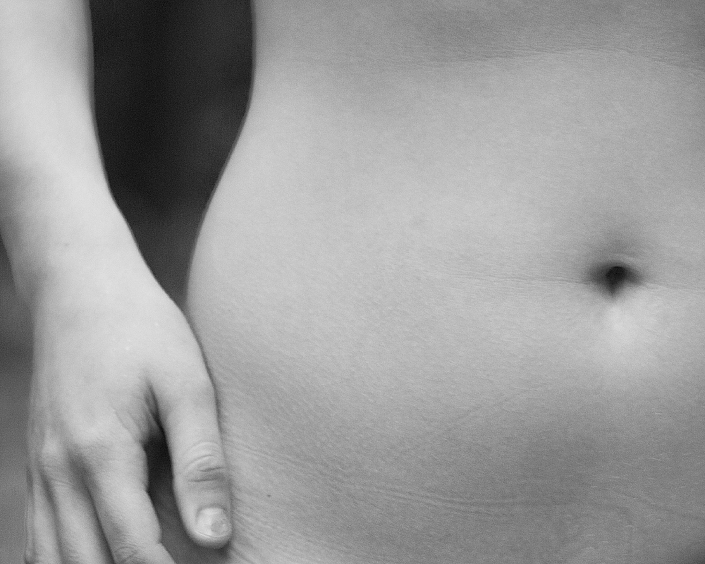 The right side of a woman’s bare stomach and right forearm are visible as she stands facing the camera.