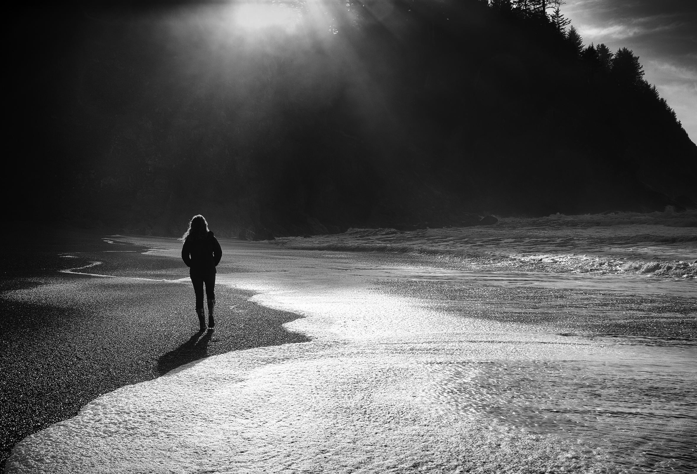 Woman walking into the morning sun on the beach. The coastline she is walking toward is hilly and tree-covered. She is shot from the back and is wearing a jacket, jeans, and boots.