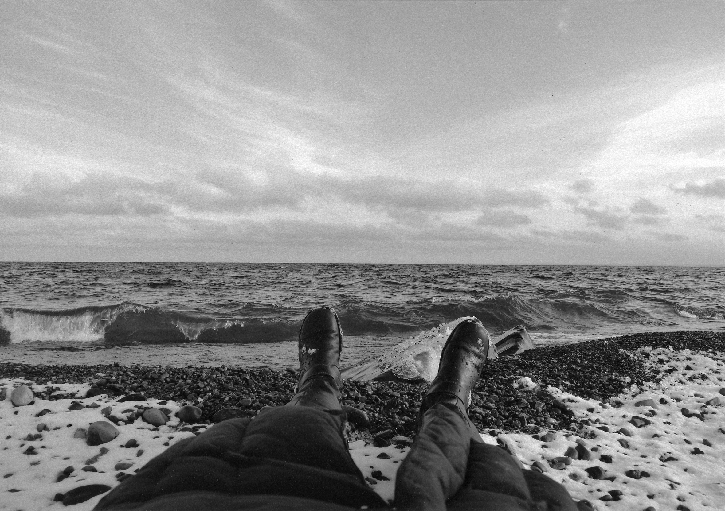 The bottom part of a person in a puffy coat and boots lying on their back on a snow-covered, black, rocky beach with their boots pointed toward the ocean.