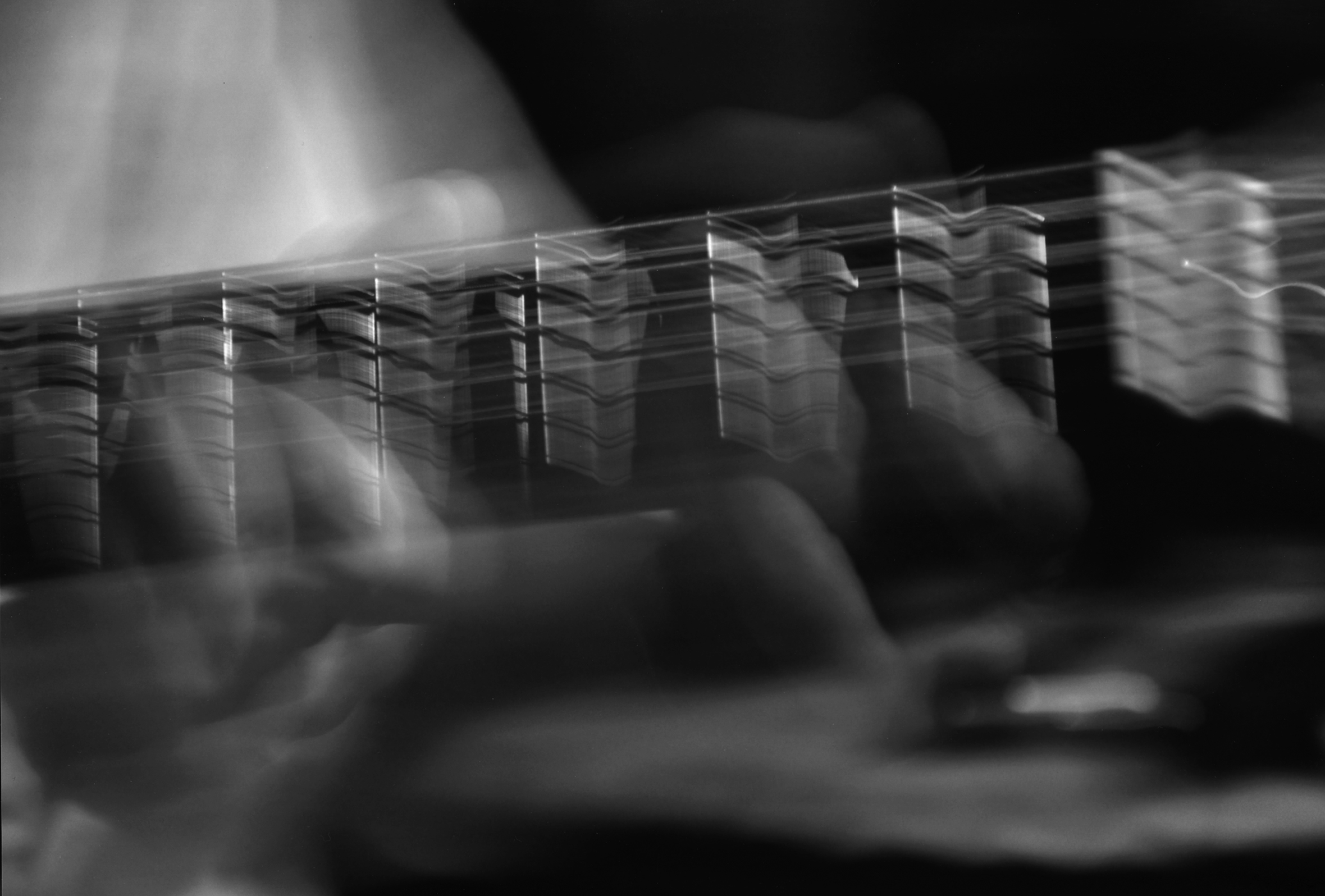 Hands play the strings on the neck of a guitar. The image has been filtered and blurred so it looks like you can see the movement of the fingers and hands while playing.