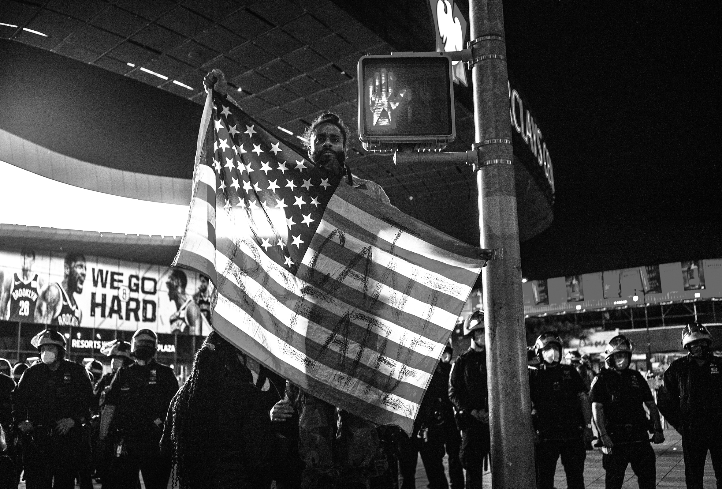 A demonstrator at a protest in New York City, in May 2020, is holding an American flag with “I can’t breathe” on it, at the same height as the crosswalk signal illuminating the do not walk hand icon. Behind the protestor is a large number of officers.