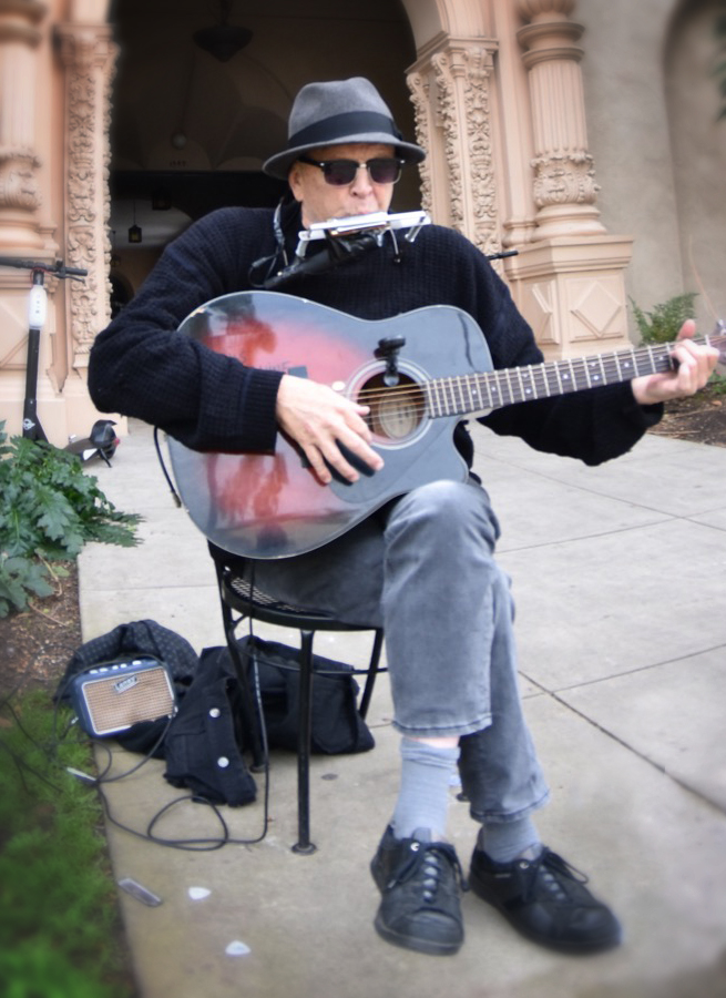 Saint James Harris Wood, wearing a hat and sunglasses, black shirt and jeans. He's playing a guitar and harmonica while sitting in front of a building.