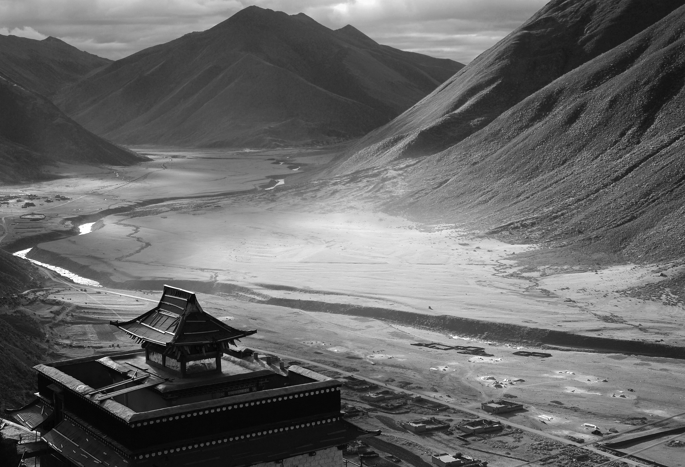 A landscape shot of a valley. Mountains are off in the distance. A river cuts through the middle. In the foreground, along the left corner is a Buddhist temple.