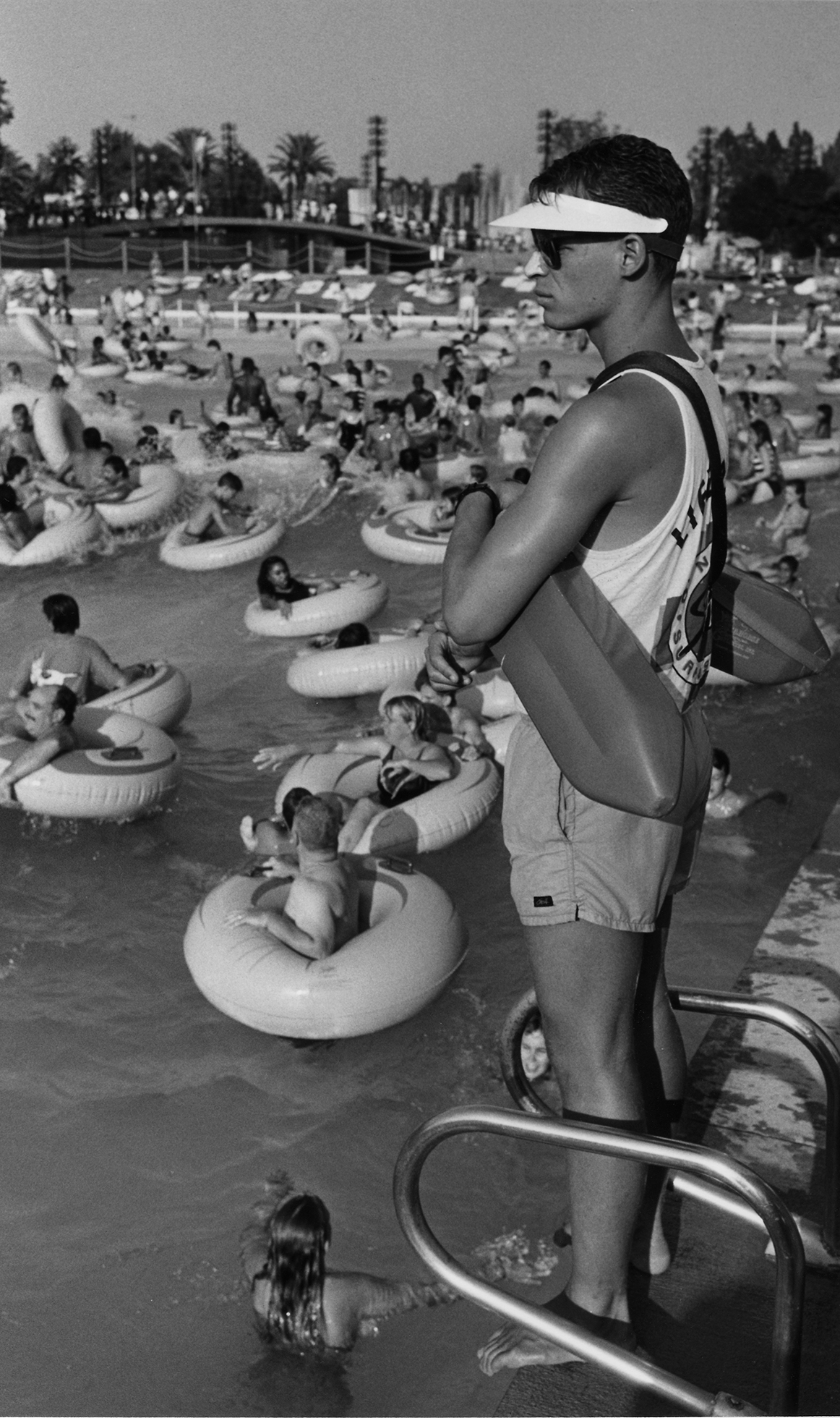 A male lifeguard wearing a visor, sunscreen on his nose, and sunglasses is standing along the edge of a pool. He’s looking off in the distance. Beyond him are clusters of people floating in innertubes.