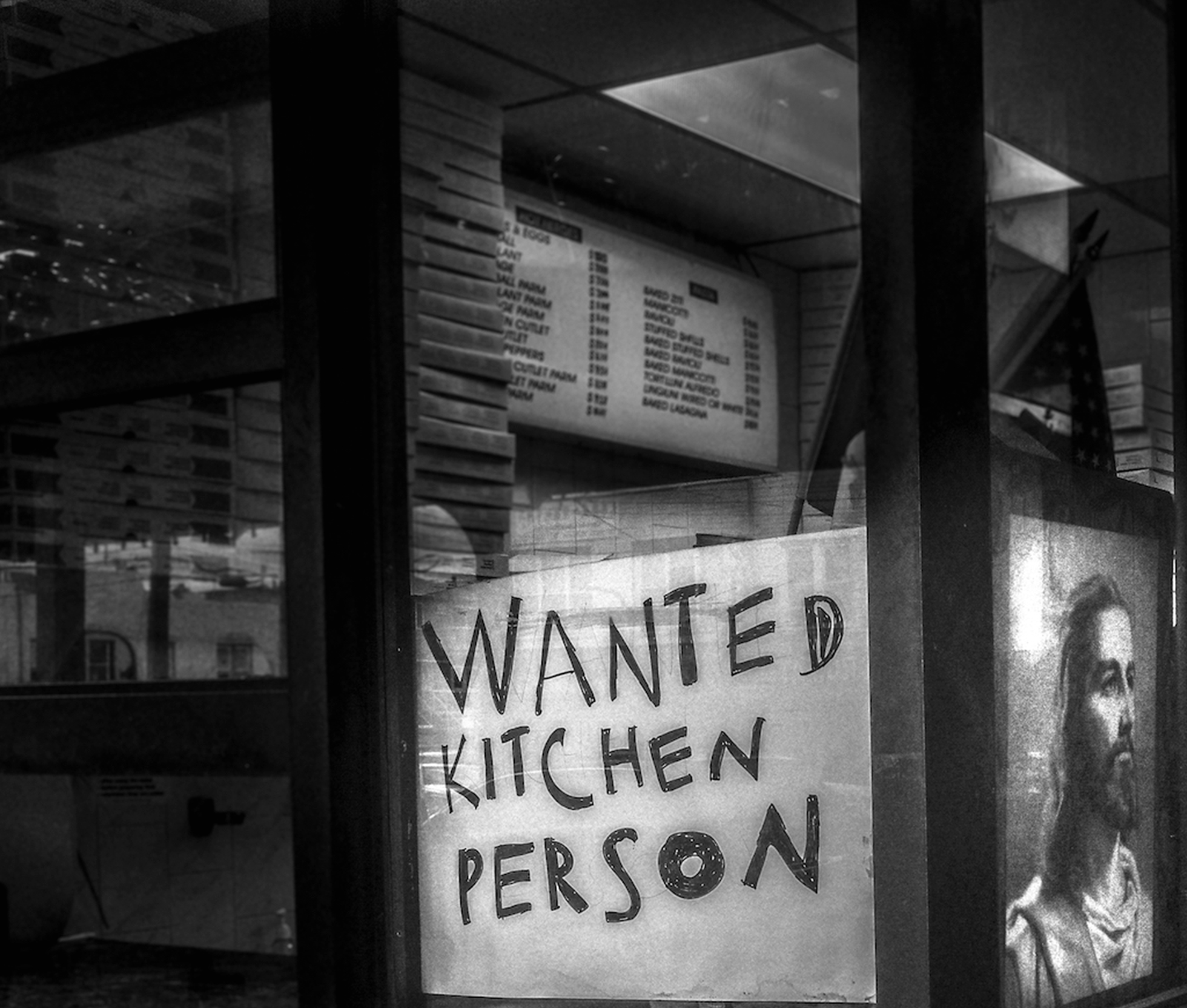 A sign that says Wanted Kitchen Person is placed in a restaurant window. In the window next to it is an image of Jesus Christ.