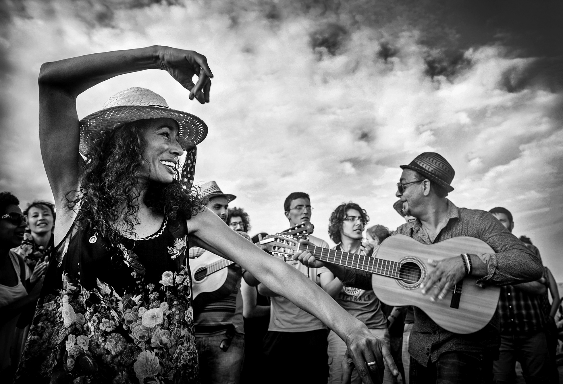 A woman on the left is in a floral shirt and straw hat and is dancing with a big smile. To the right is a man playing a guitar and he's looking over at another man playing a guitar. A group of joyous people surround them.