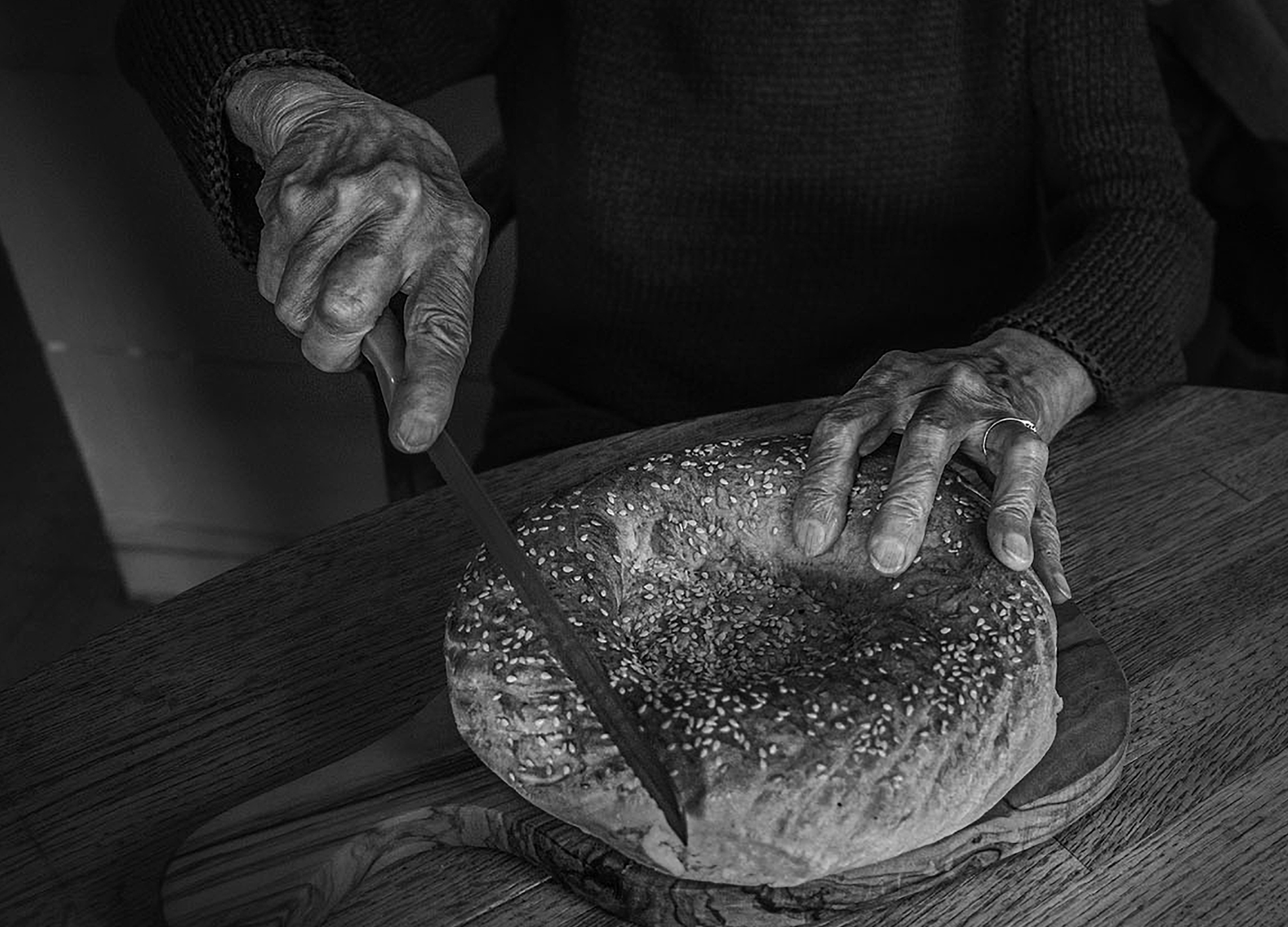 A close-up of an old person’s hands as they cut into freshly baked bread that’s been sprinkled with seeds.