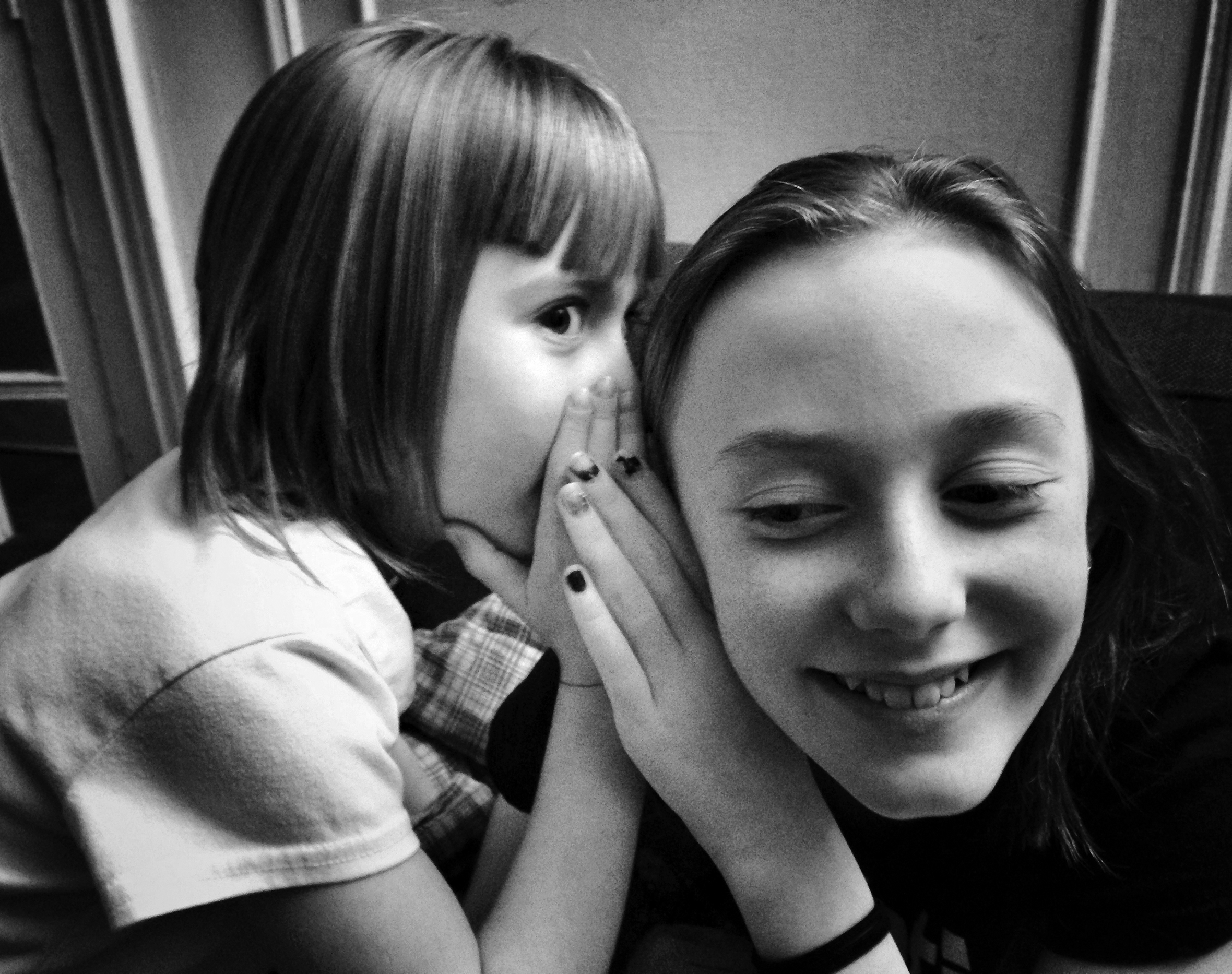 A young girl whispers in a teen girl’s ear. The teen girl listens closely and smiles.