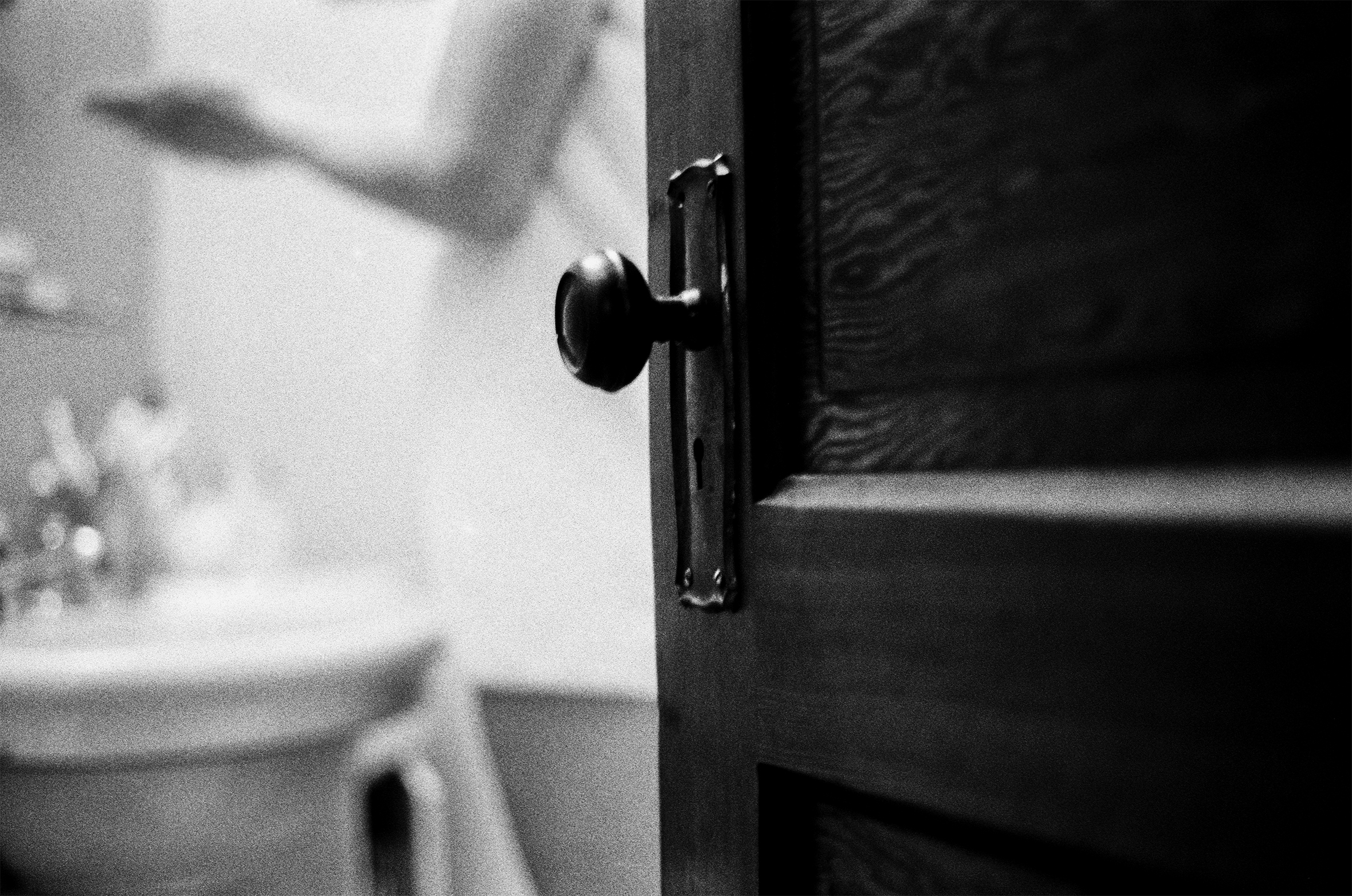 A bathroom door is cracked open. Out of focus in the background, a woman stands in front of the sink.