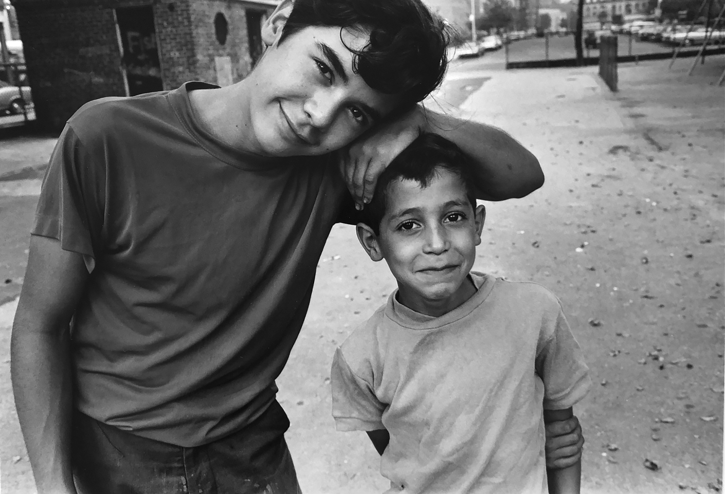 Two boys stand and smile at the camera. The older boy rests his arm on the top of the younger boy and leans his whole body toward the younger one.