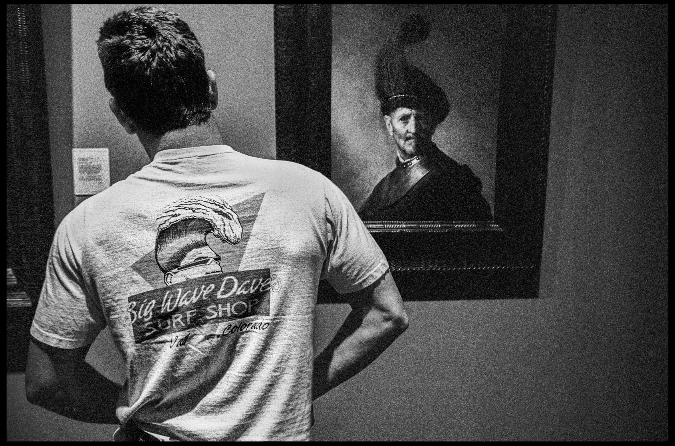 A man wearing a Big Wave Dave’s Surf Shop T-shirt is standing with his hands at his waist as he looks upon Rembrandt van Rijn’s An Old Man in Military Costume painting at a museum.