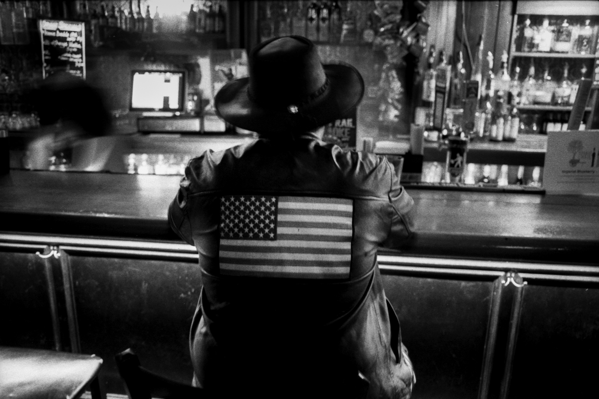 A person is sitting at a bar wearing a leather jacket with the American flag across the back and a Western-like hat.