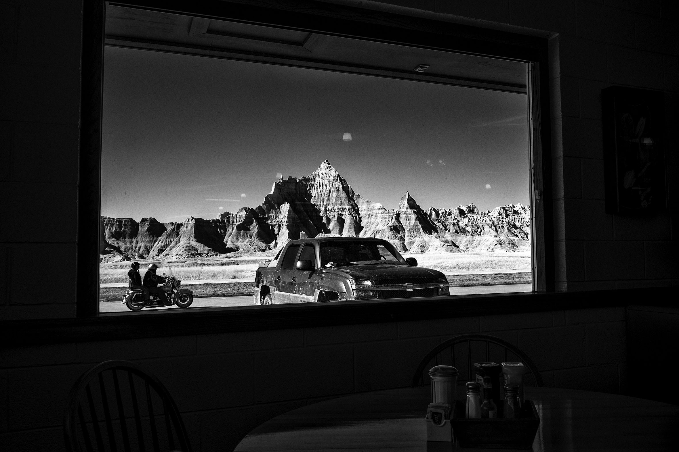 Scenery outside of a diner window of desert cliffs and plateaus in the distance. A truck is parked in front of the window and two people on a motorcycle are riding through the parking lot.