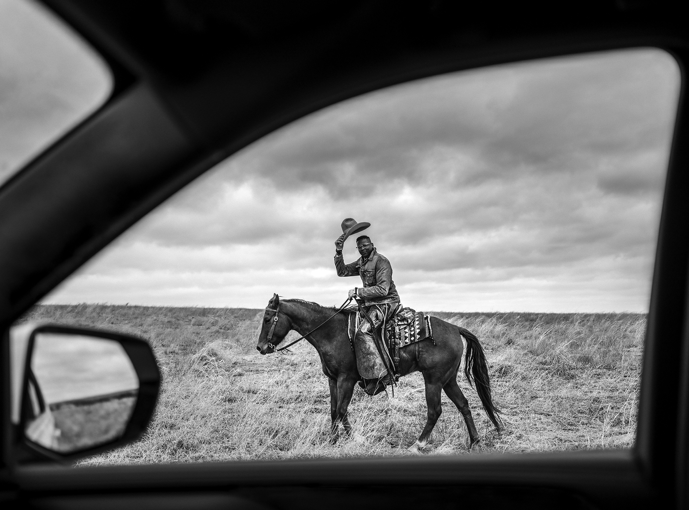 A Black cowboy on his horse. He’s tipping his hat to the person holding the camera. The image is framed by a car interior.