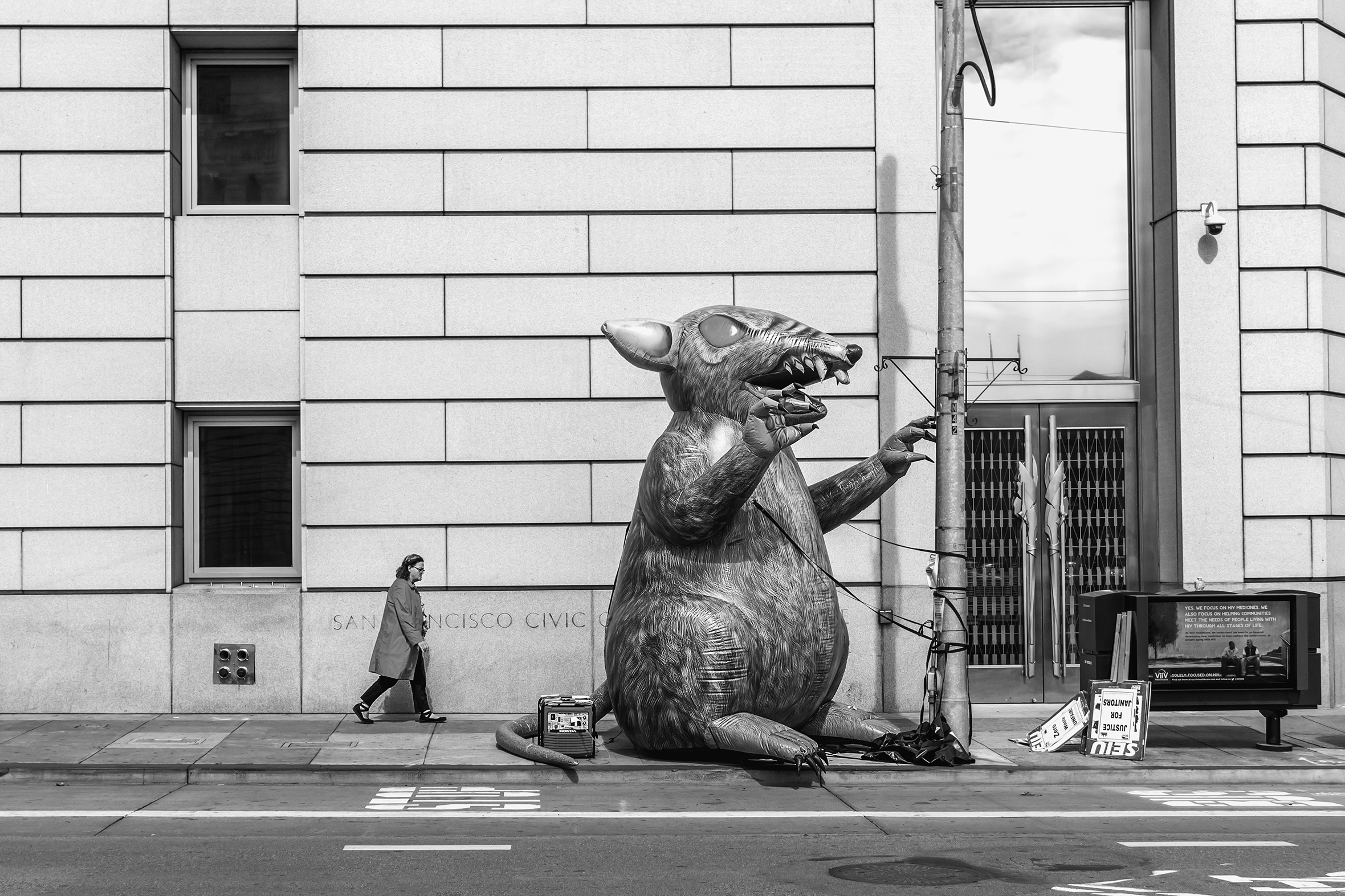 A one-story tall, violent-looking, inflatable rat is tethered to a street pole outside the San Francisco Civic Center. Next to the rat are SEIU picket signs that mention Justice for Janitors. A woman walks by the rat.