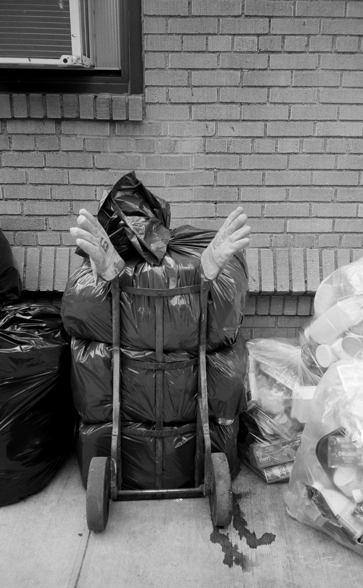 A dolly holding three trash bags is in front of a brick building. The gathered part of the topmost bag is sticking up and atop each dolly handle is a glove so that it looks like the front of a person whose head is tilted to the right.