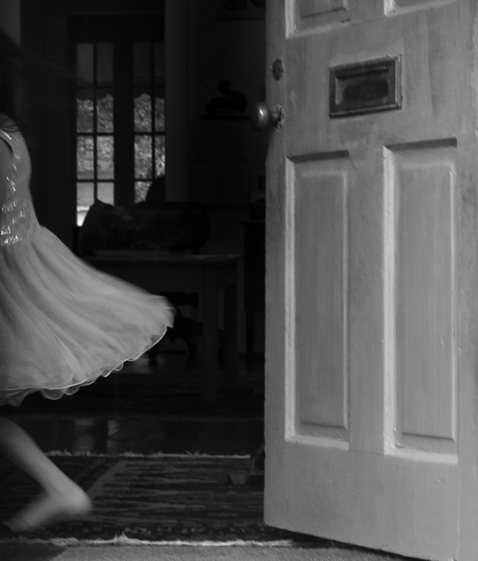 A barefoot girl has dashed into a home through the front door leaving it open on the right. She is moving to the left and a glimpse of her white dress with ruffles on the bottom and sparkles on the top back is captured as she disappears inside.