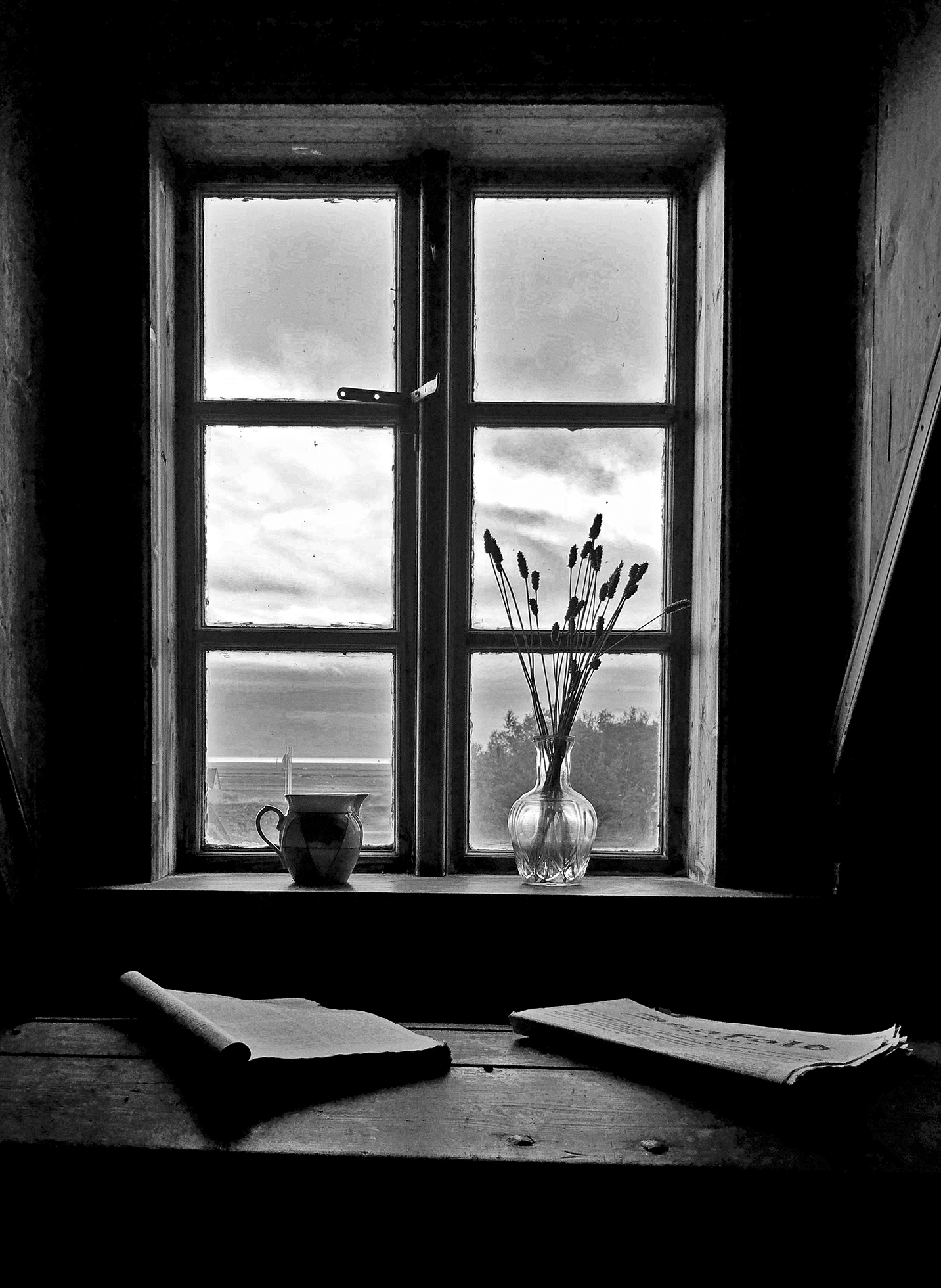 A clear vase with tall, slender flowers and a small pitcher sit on a windowsill. The lighting illuminates just enough of the interior to see an opened book and a folded newspaper on a bench in front of the windowsill.