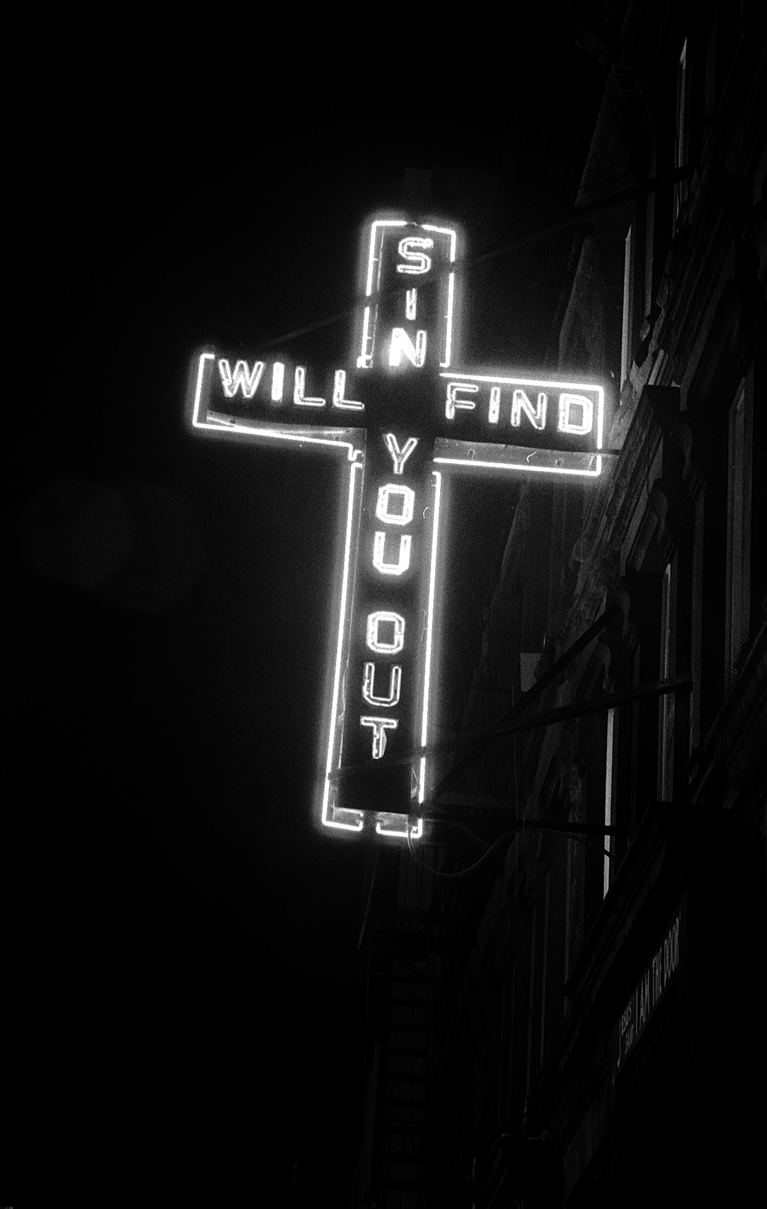 A large neon sign of a cross attached to the side of a multistory building photographed at night that reads SIN WILL FIND YOU OUT.