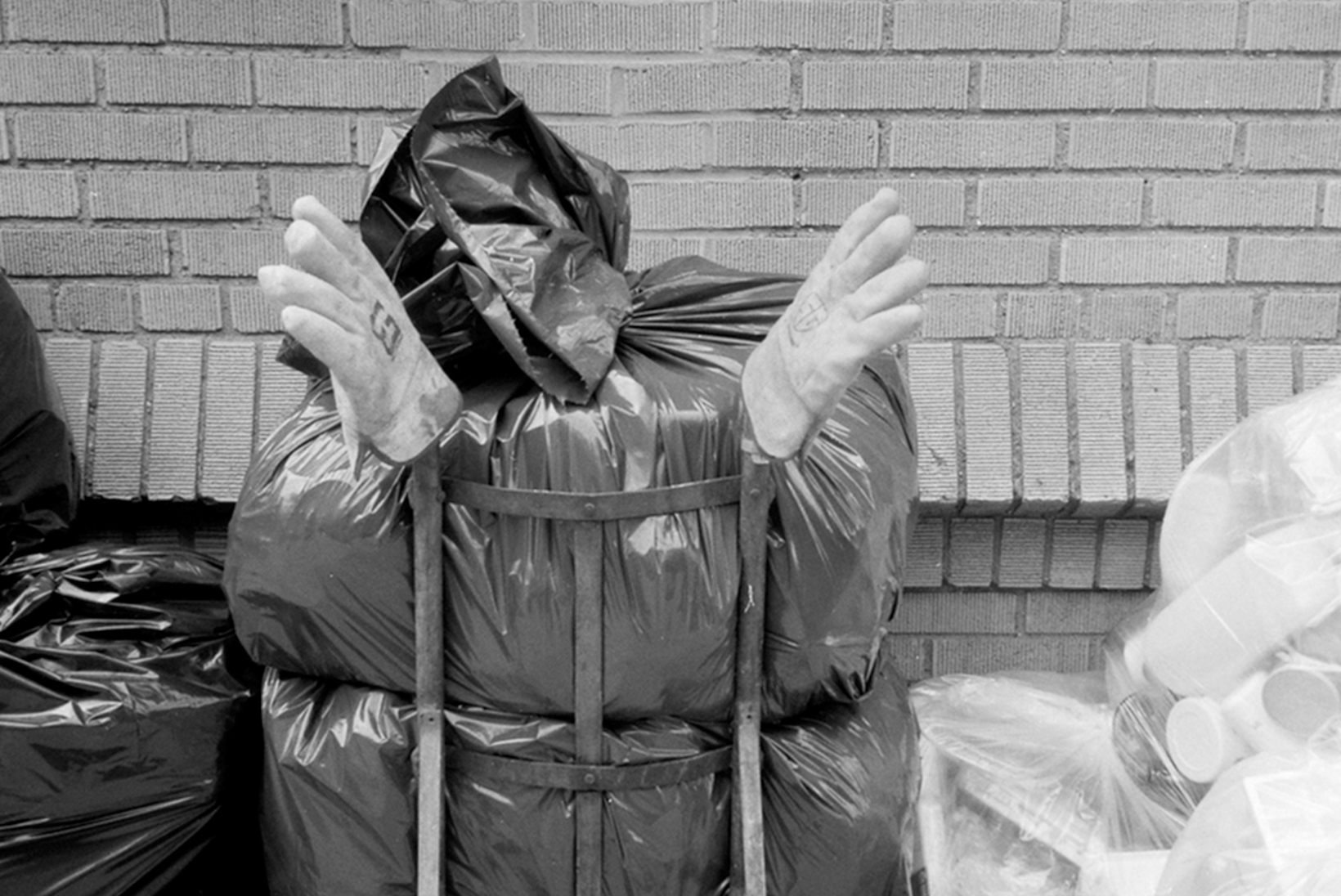 A dolly holding three trash bags is in front of a brick building. The gathered part of the topmost bag is sticking up and atop each dolly handle is a glove so that it looks like the front of a person whose head is tilted to the right.
