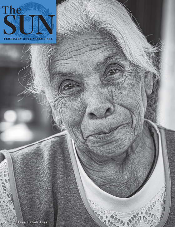 February 2022 cover of The Sun. A close-up of an older woman with gray hair who has a closed-mouth smile and kind eyes and is wearing a white shirt and a vest.