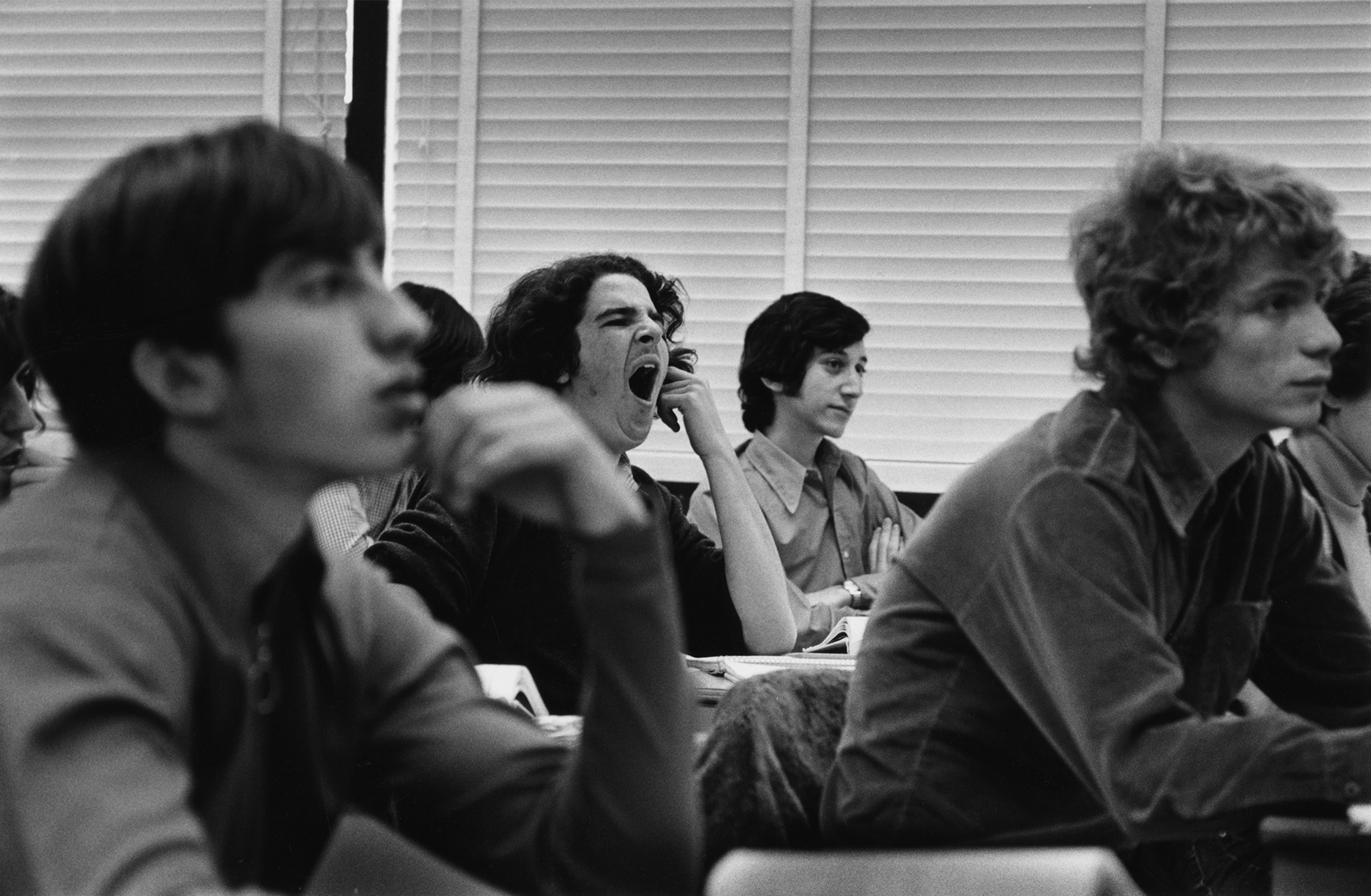 Teenage boys are sitting in a classroom and paying attention to something in front of them off camera, except for one boy who is yawning. From the hairstyles it looks like the early 1970s.