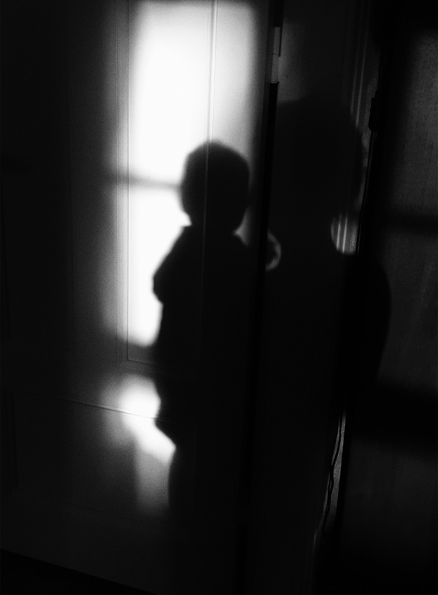 The silhouette of a woman standing and holding a small child seen from behind. The image is dark, but there is light from a large window in front of the woman and child.