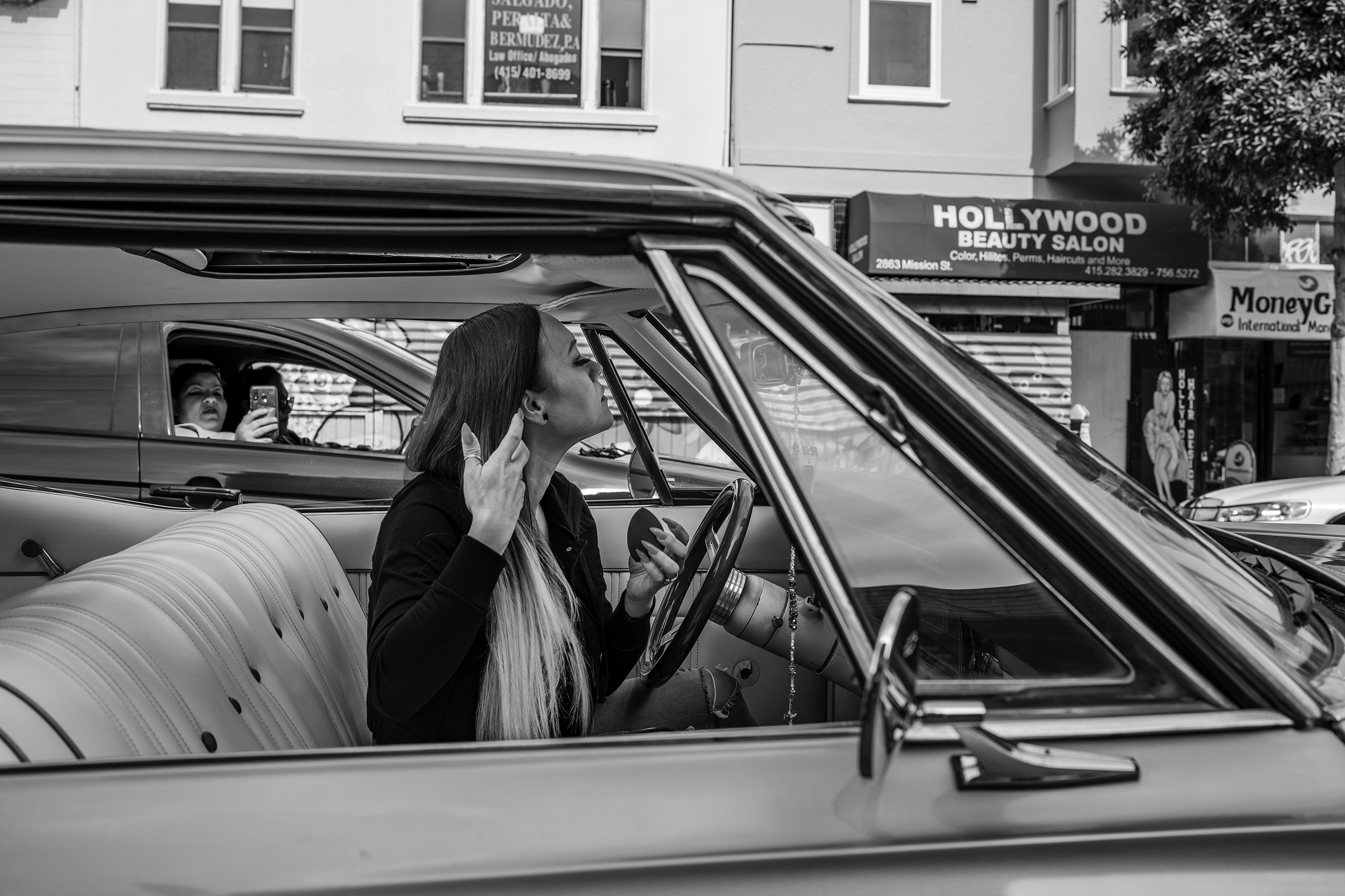 A woman is sitting in a car with a front bench seat and looking in the rearview mirror at her long, straight, ombré hair as a woman in a passing car is taking her photo with a cell phone. Across the street is a sign for the Hollywood Beauty Salon.