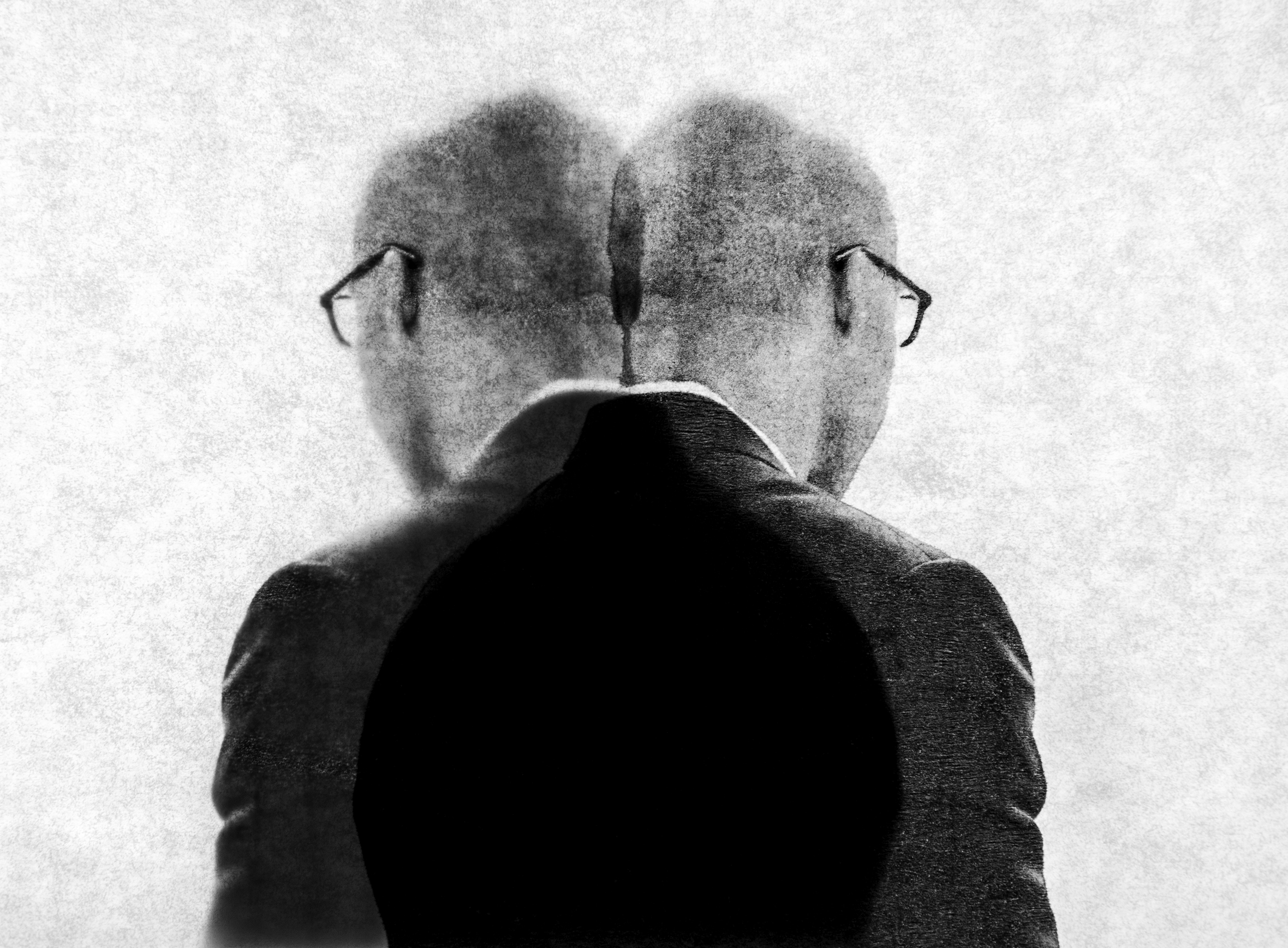 A man with close-cropped hair and black-framed glasses wearing a white shirt and dark suit coat is seen from the back. The man’s image has been duplicated and placed together so that there is one trunk but two heads looking in opposite directions.
