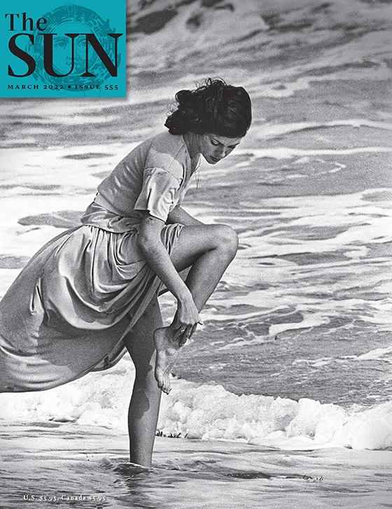 March 2022 cover of The Sun. A woman with brown hair in a short-sleeved, knee-length dress is standing in the ocean surf up to her ankles. She is standing on one leg while the other is lifted and bent at the knee as she bends over and touches her ankle.