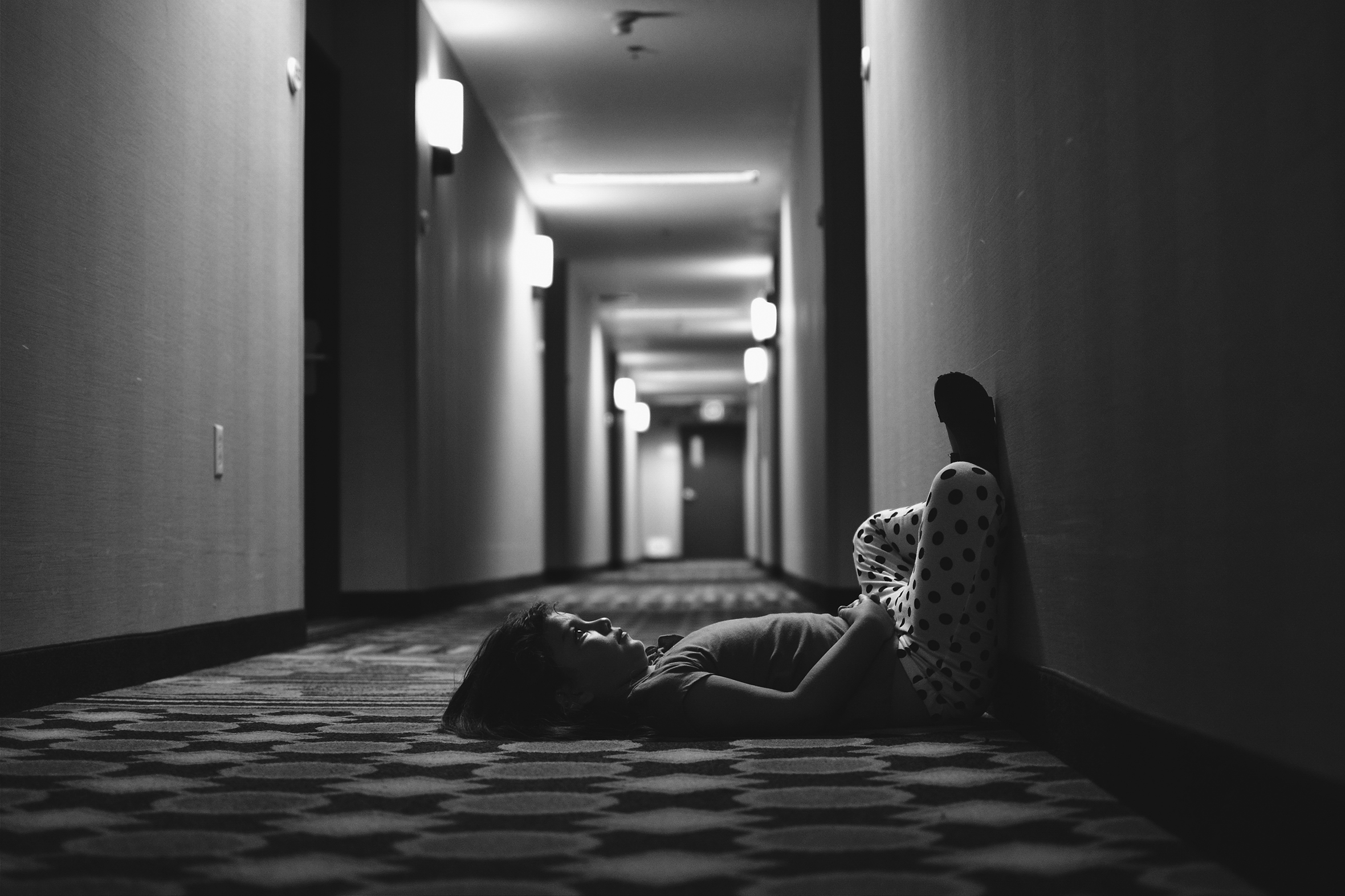 A young girl is laying on the carpet in a long apartment hallway facing the wall with her buttocks against the wall and her legs up the wall. She is staring at the wall with her hands across her stomach.