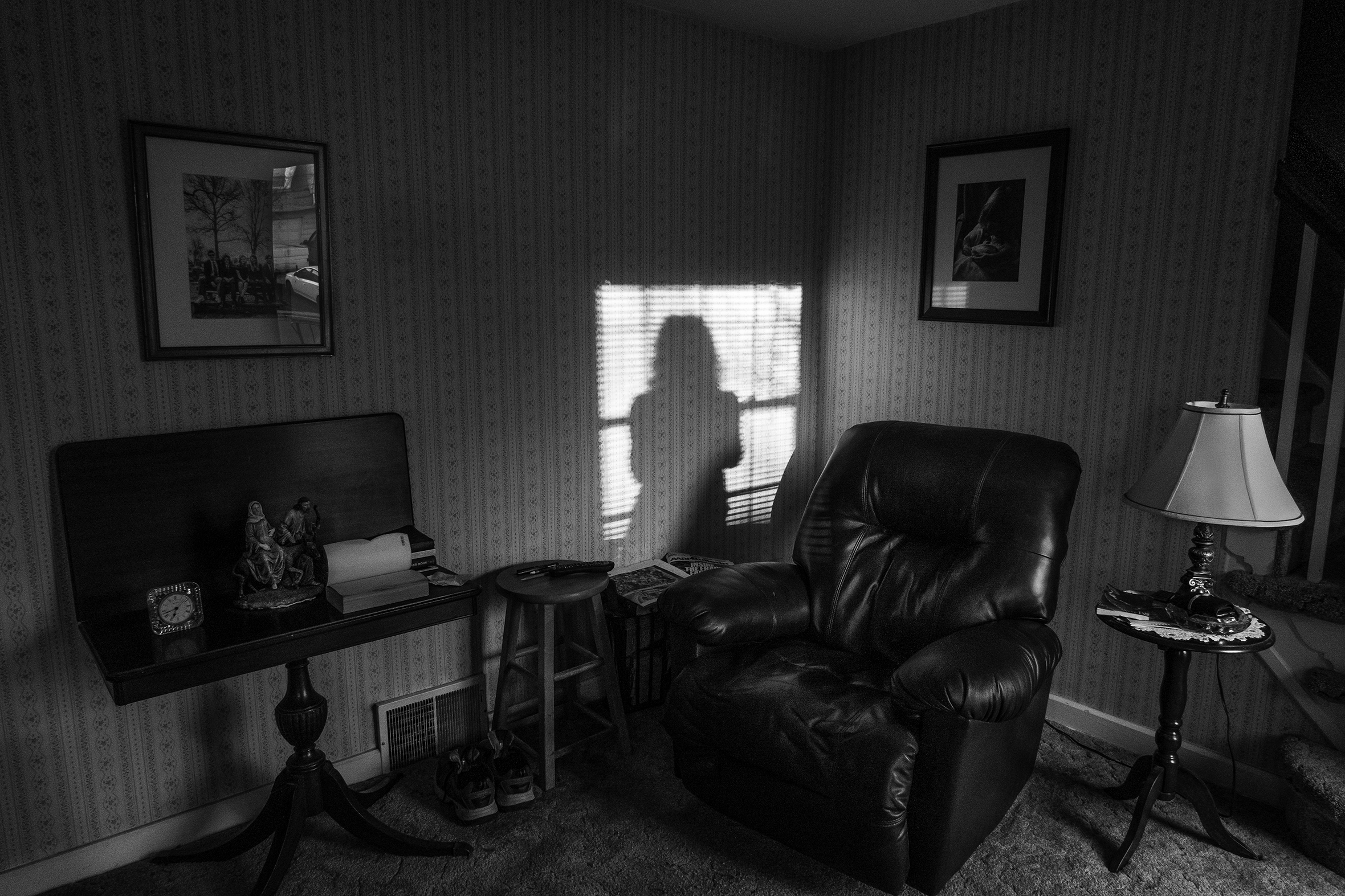 A living room domiated by an empty leather recliner. A woman stands in front of a window looking out.