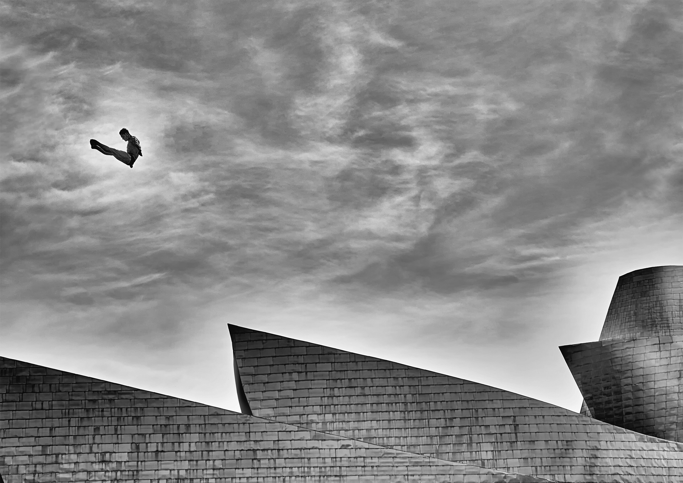 The top part of three angular roofs staggered behind each other against a backdrop of swirling clouds. In a small bright spot in the sky is a man in a speedo seen mid jackknife dive. He is rotated so that his toes are pointed up to the right.