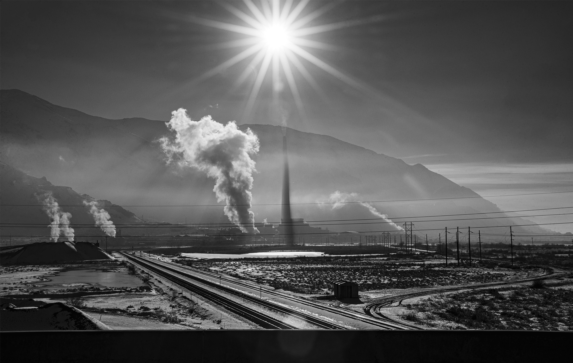 A wide shot of a desolate industrial landscape with smoke billowing from several smokestacks. There is a sunburst at the top middle of the image that is dulled by the pollution in the air.