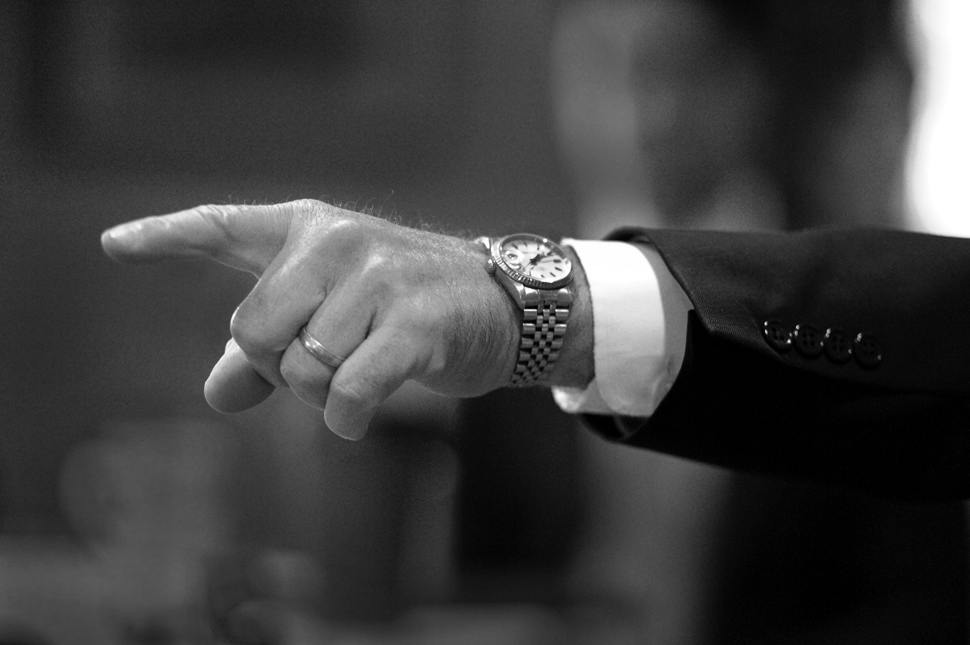 A man’s left forearm is extended as he points his index finger. A wedding band and watch face are visible, as is the sleeve of a suit coat with a white shirt cuff peeking out.
