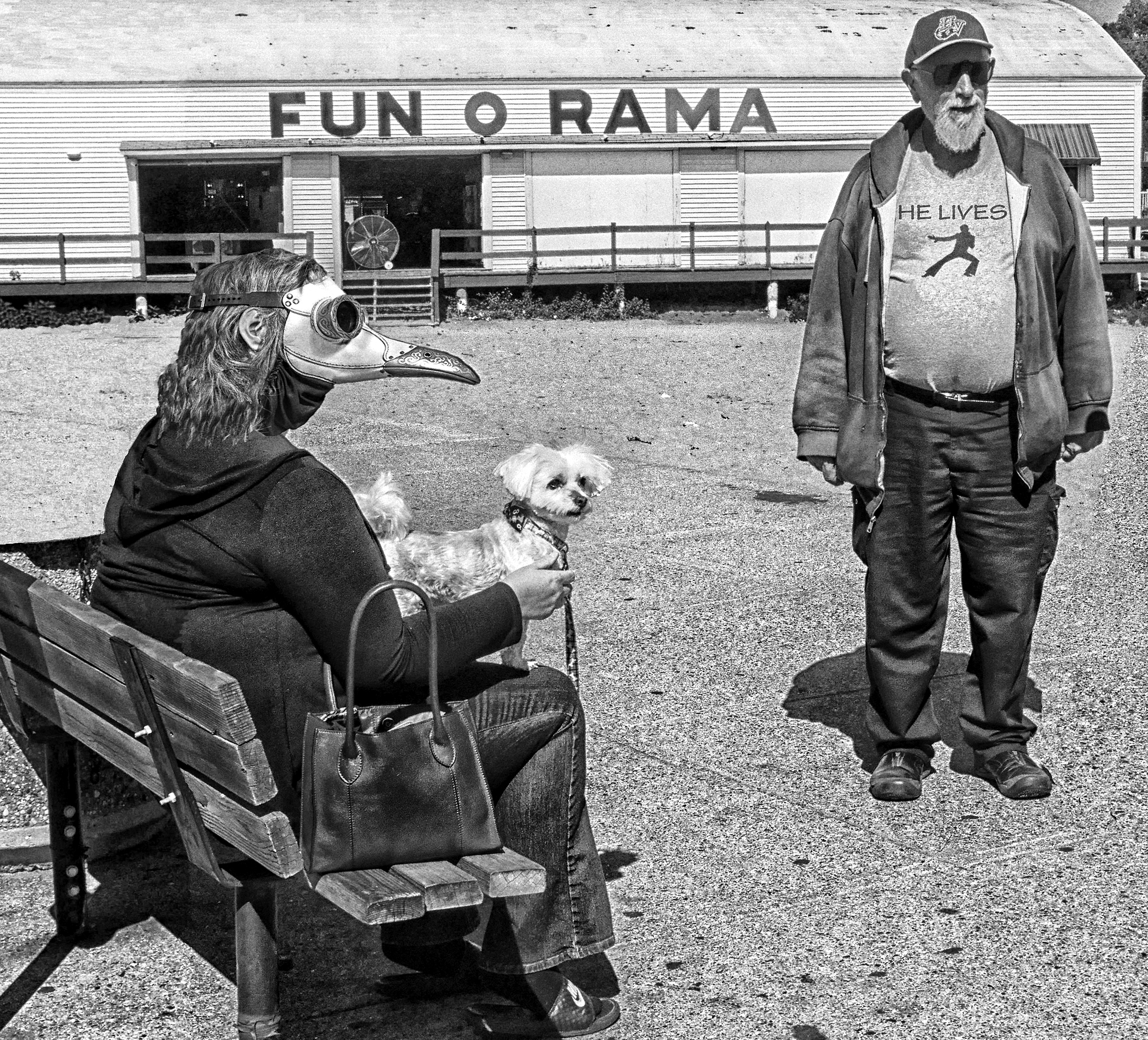 An old woman is sitting on a bench wearing a white plague-doctor mask with a Maltipoo on her lap. Standing near her is an old man wearing a T-shirt with “He Lives” and a silhouette of a jumpsuited Elvis. Behind them is a building with “Fun O Rama” on it.