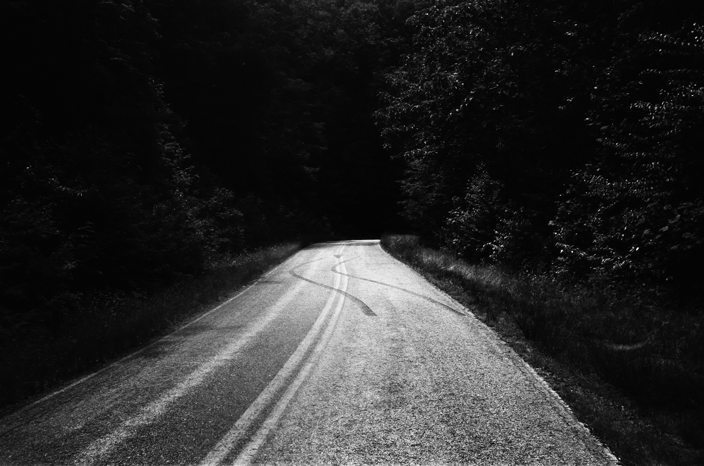A short length of a two-lane road in the woods at night curving off to the right in the near distance. There are skid marks from the right lane into the left and then back to right.