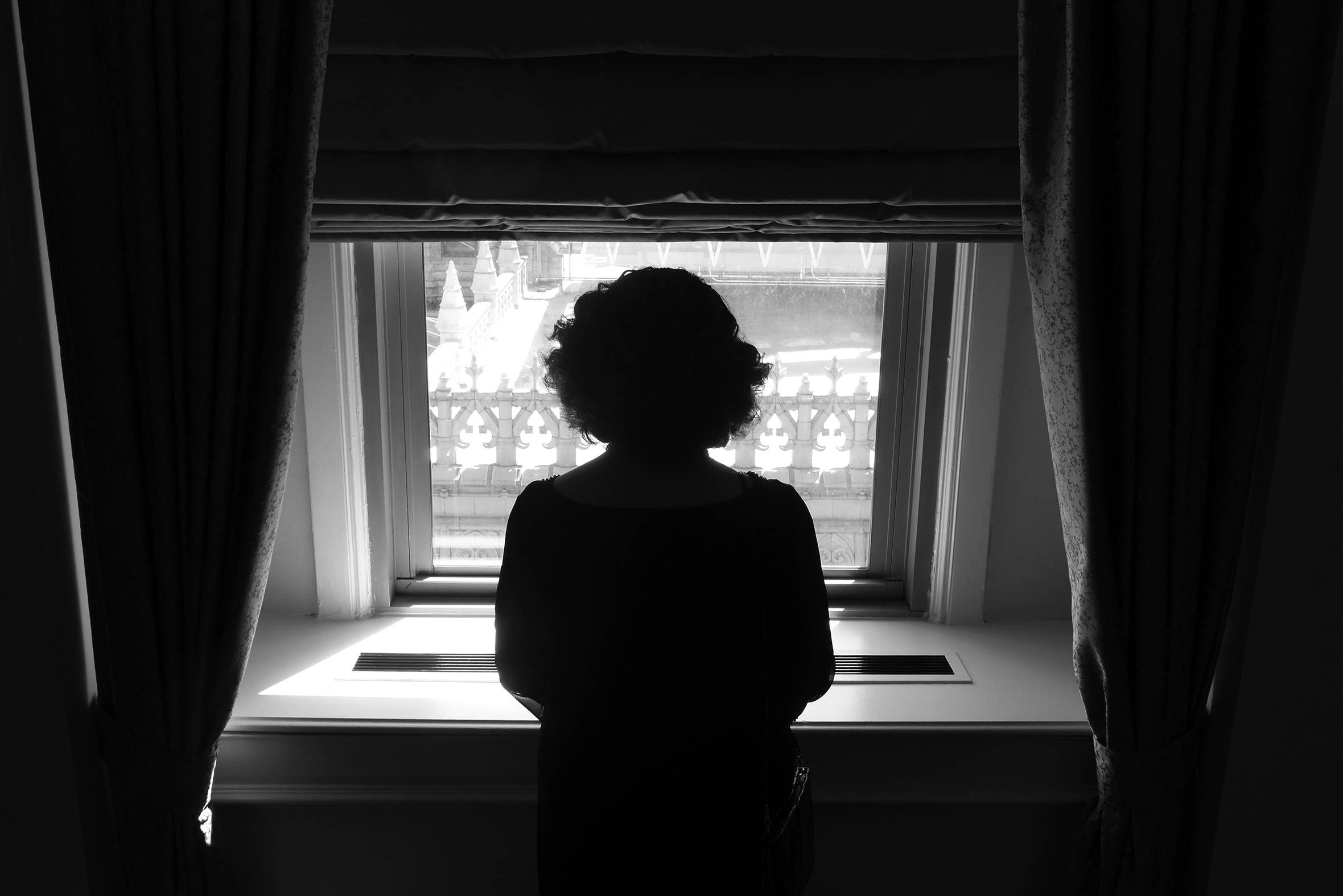 The back view of a woman in a dark room looking out a window In the daytime. The window has heavy curtains pulled to the sides and a shade on top. The windowsill is white and very deep, with a long radiator cover across the center.