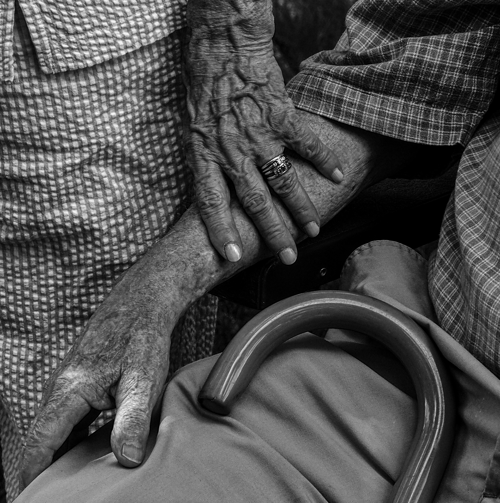 An old woman’s hand rests upon an old man’s forearm as she stands above him. He is seated with a cane in his lap.