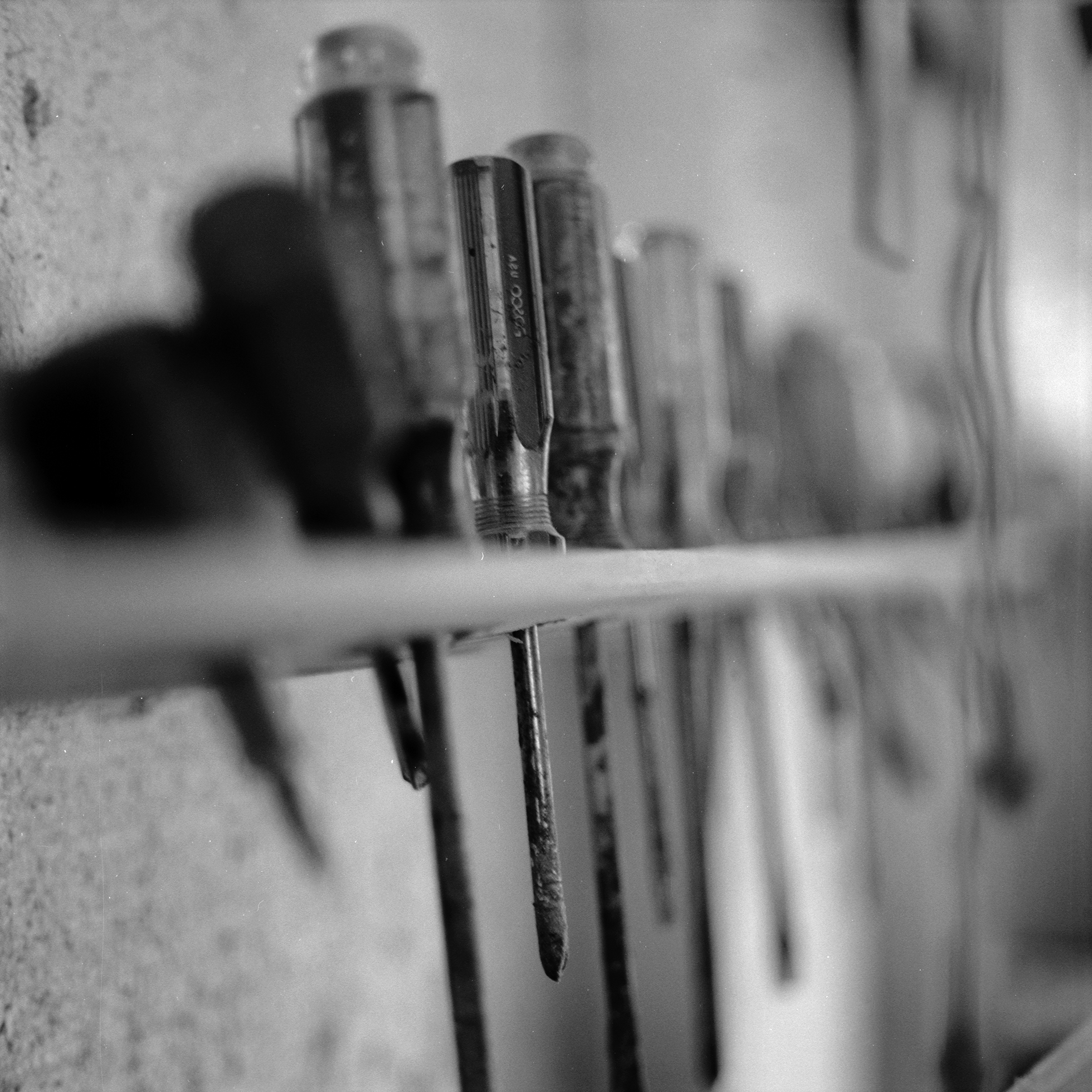 Well-used tools, each in its own hole, are stored along a narrow wooden strip attached to a wall. The screwdrivers in the foreground are in focus, while the rest of the tools are blurred.