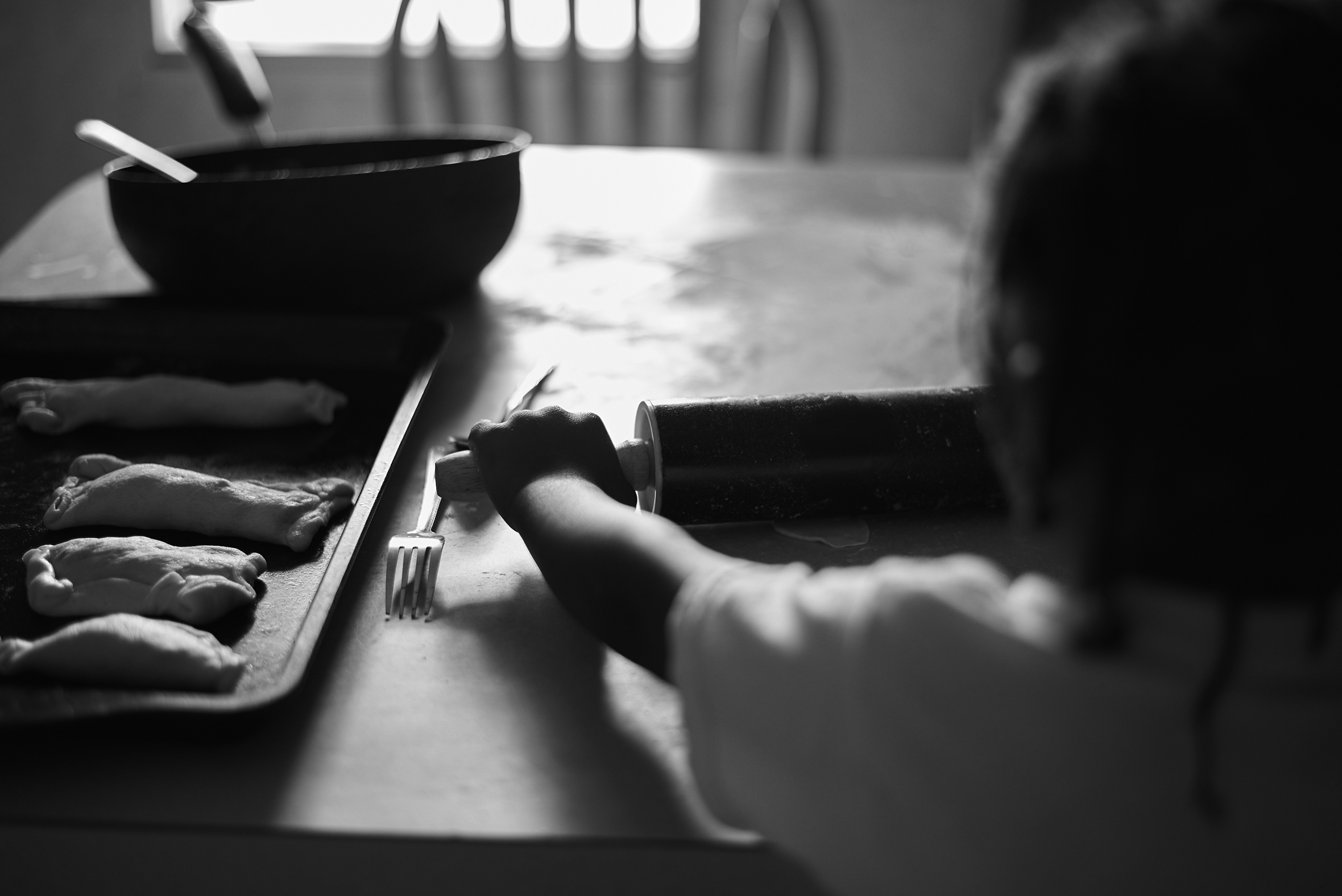 A young girl stands at a floured kitchen table rolling out a small amount of dough. A cookie tray with rectangular, stuffed dough creations on it is on the table to her left, and a bowl with a utensil handle sticking out is beyond the cookie tray.